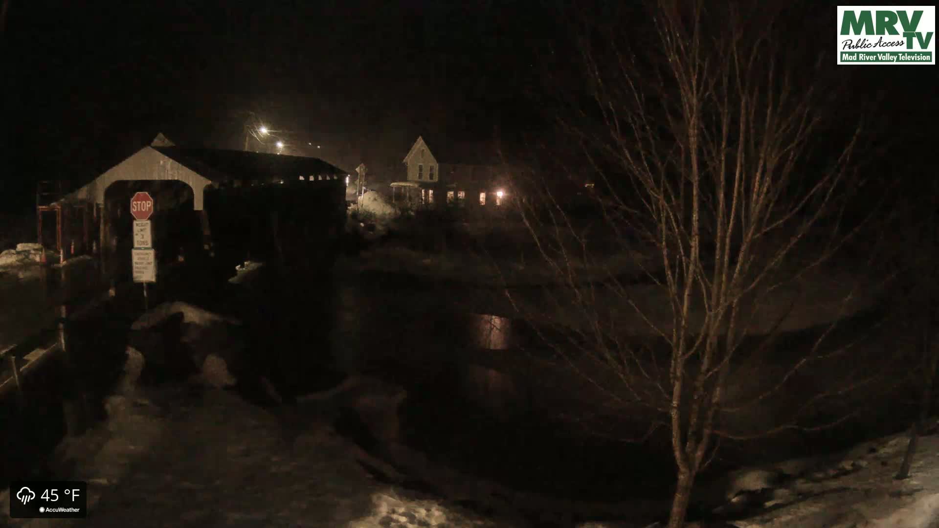 A nighttime scene shows a covered bridge, a house, and a tree, with snow on the ground and a dark body of water nearby under a dark, possibly cloudy sky.