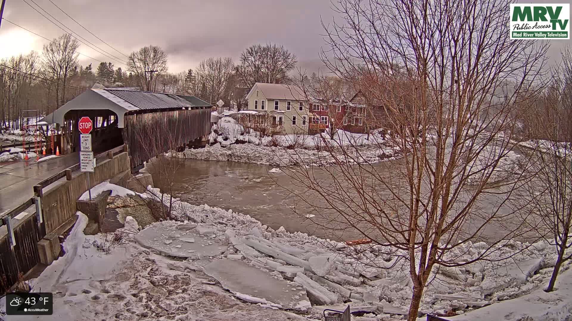 A covered bridge spans a river partially covered with broken ice floes, under an overcast sky, with snow on the ground and buildings visible in the background.