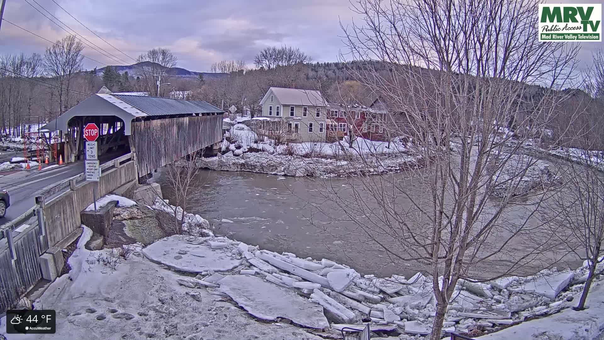 A covered bridge spans a partially frozen river, with snow-covered banks and bare winter trees in the background, under an overcast sky.