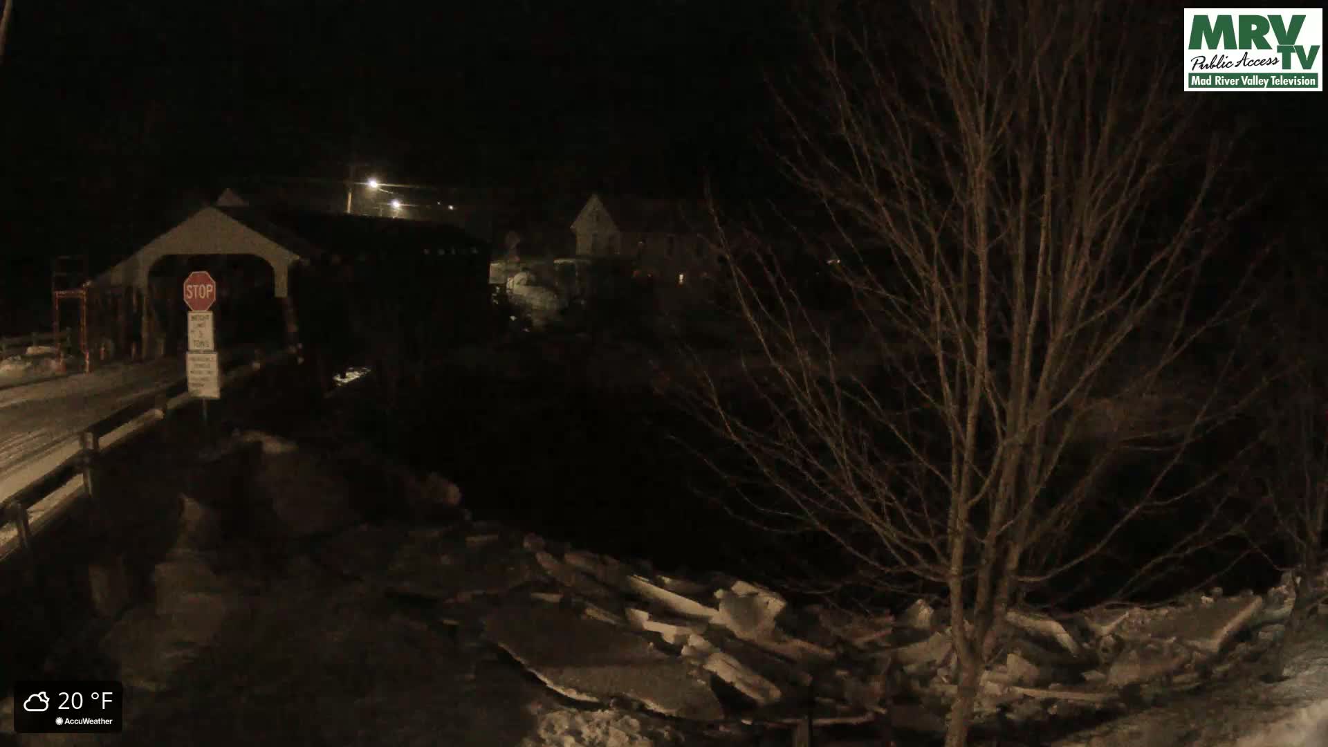 A snow-covered, nighttime view of a covered bridge with a stop sign, partially obscured by bare trees and icy debris in the foreground, at 20°F.