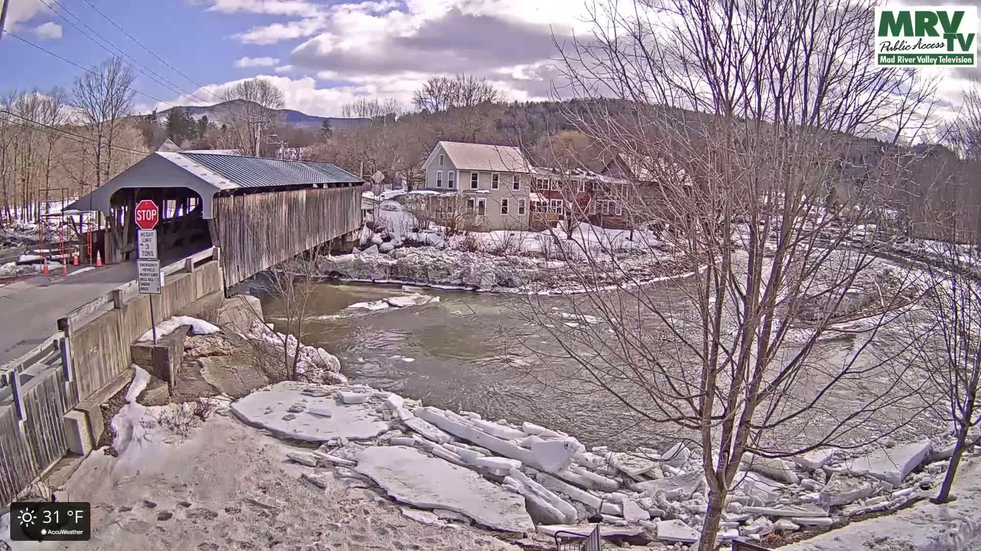 A covered bridge spans a river partially covered in broken ice floes, with snow-covered banks and houses in the background under a partly cloudy sky.