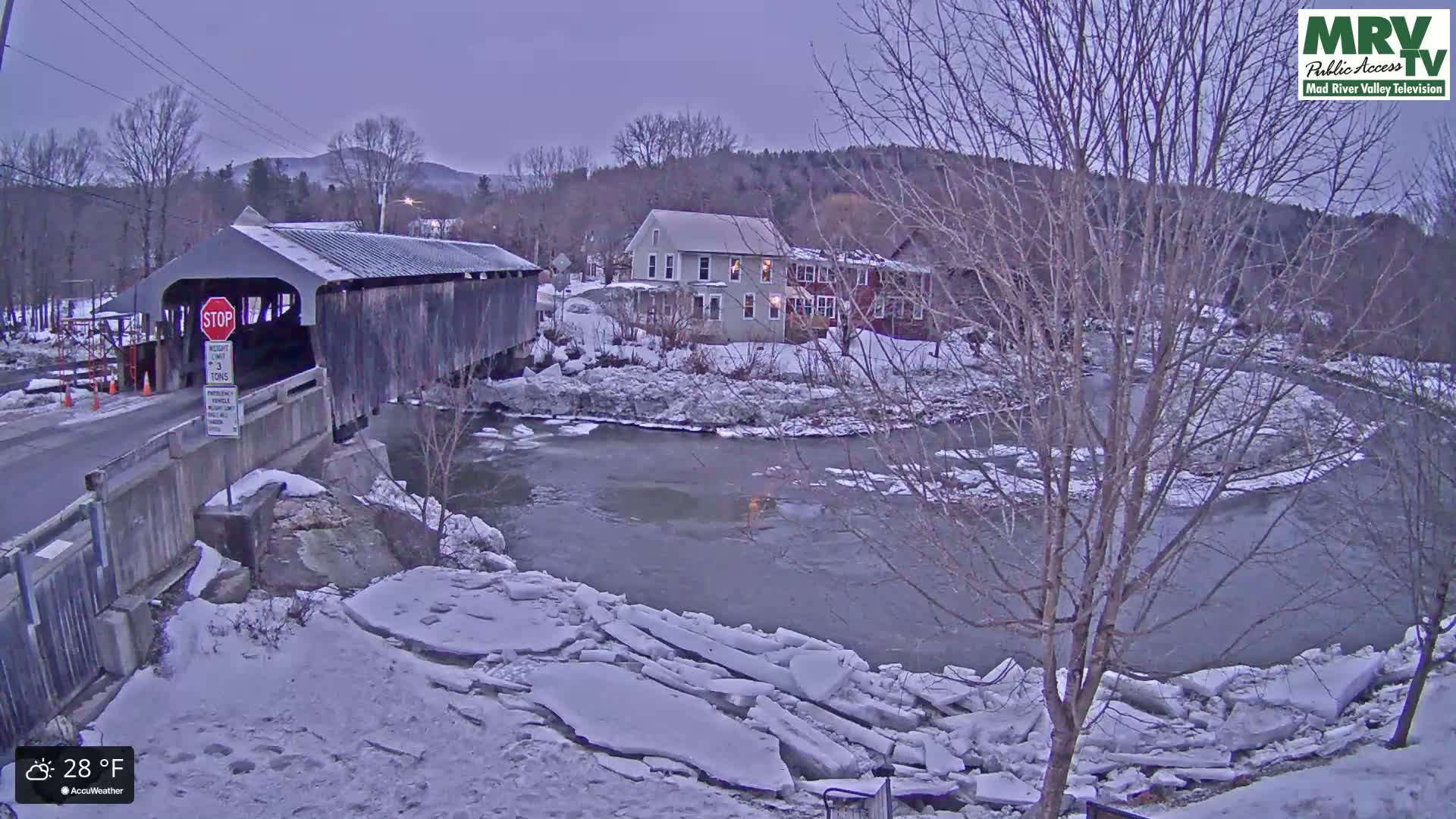 A covered bridge stands over a partially ice-covered river on a cold, twilight evening.