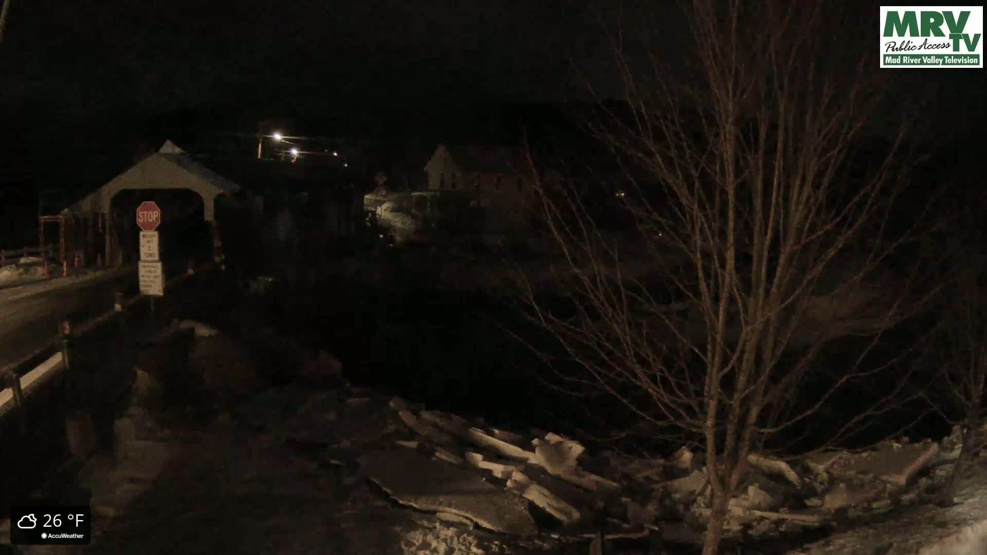 A nighttime view of a covered bridge and a snow-covered riverbank under a dark sky with a temperature of 26°F.