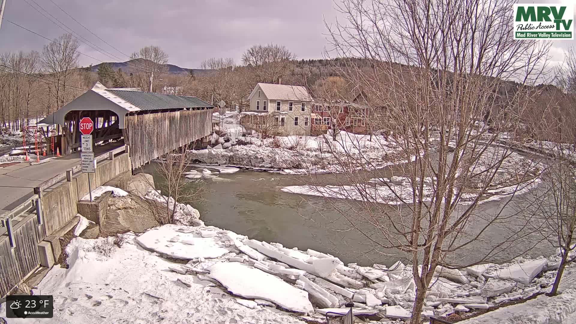 A covered bridge spans a partially frozen river, with broken ice floes along the banks, under a partly cloudy sky.