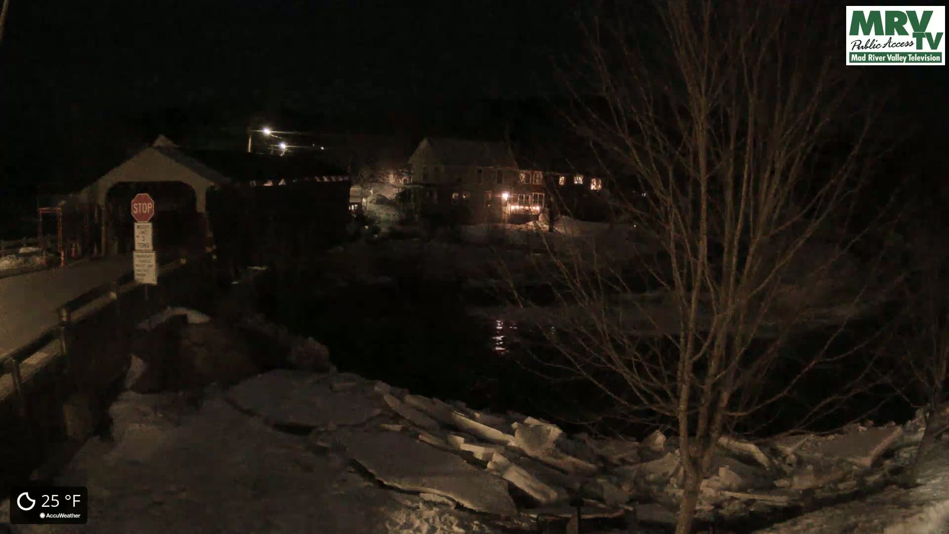 A nighttime view of a covered bridge and buildings along a partially frozen river at 25°F.