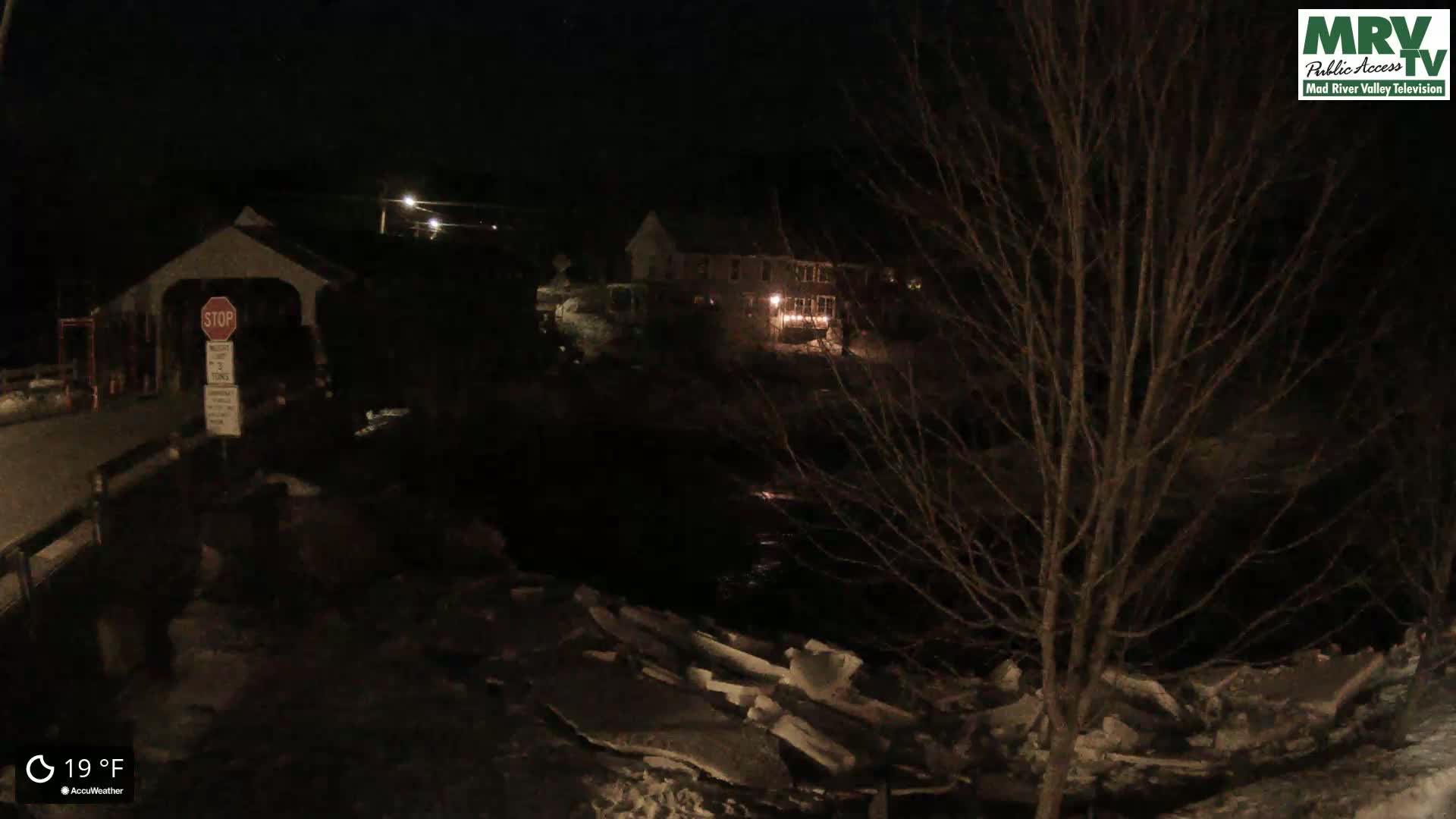 A nighttime, snow-covered scene shows a covered bridge, a nearby building with lit windows, and a dark river with ice along its banks, under a clear sky at 19°F.