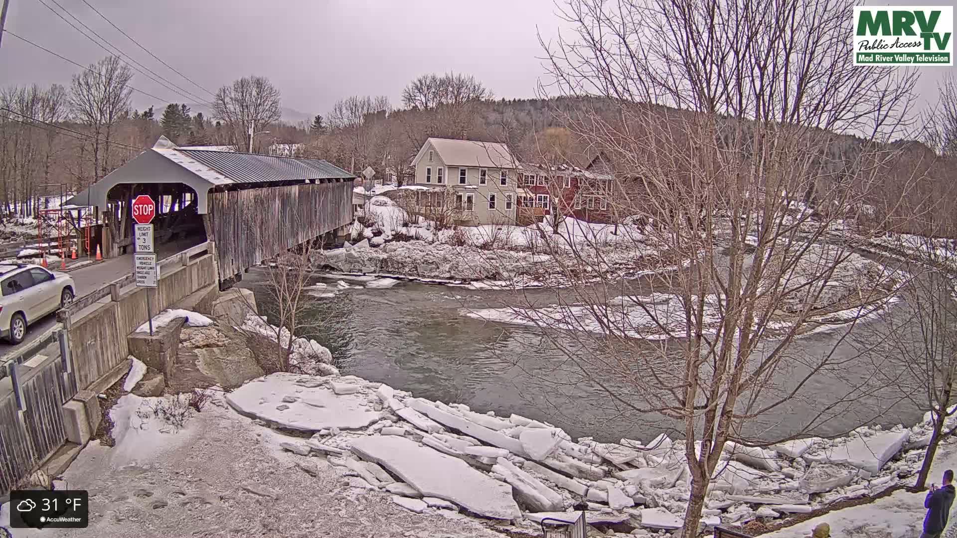 A covered bridge spans a partially ice-covered river on a cool, cloudy day, with houses and bare trees lining the riverbank.