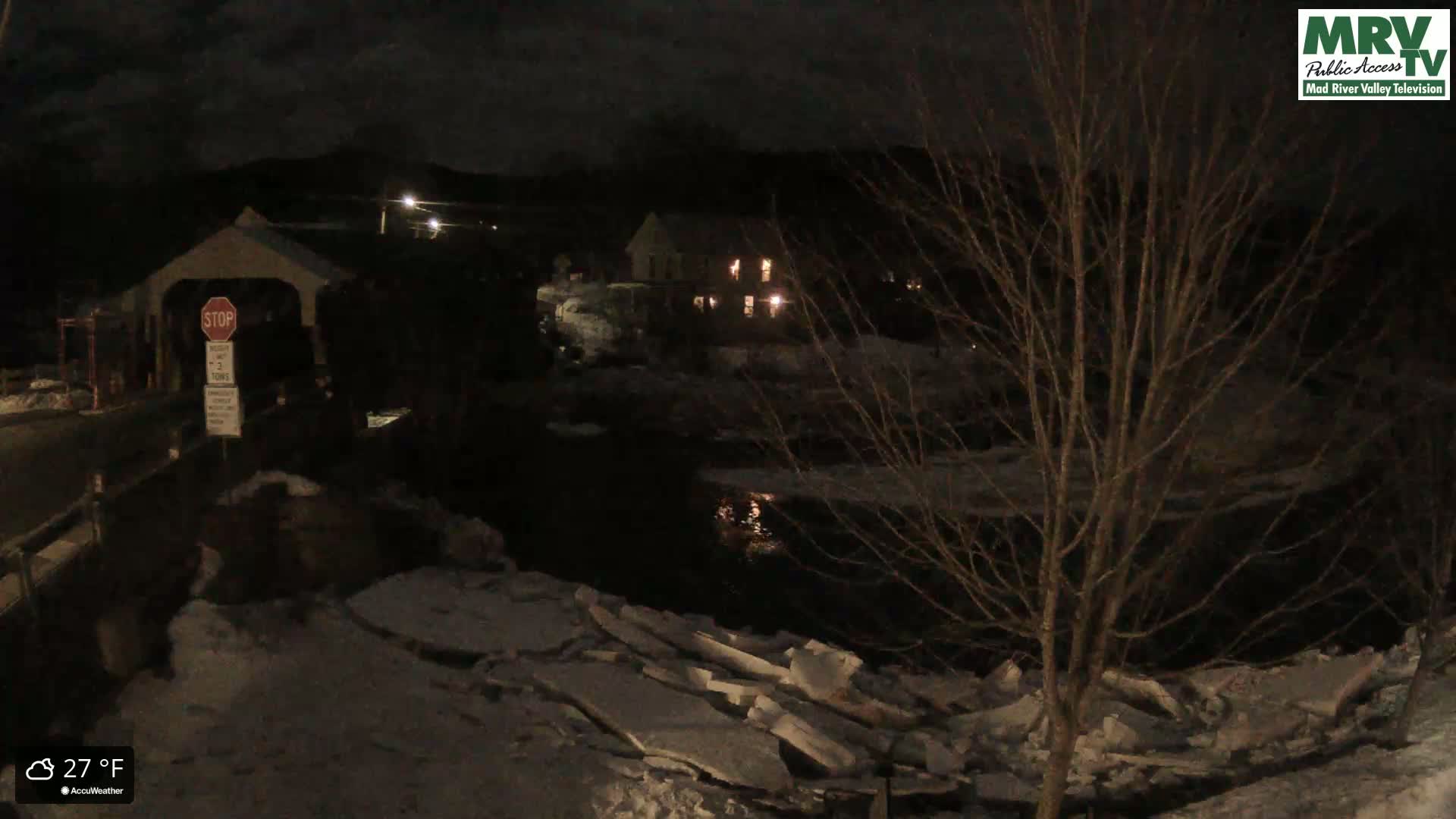A nighttime view of a covered bridge and a building near a partially ice-covered river under a dark sky.