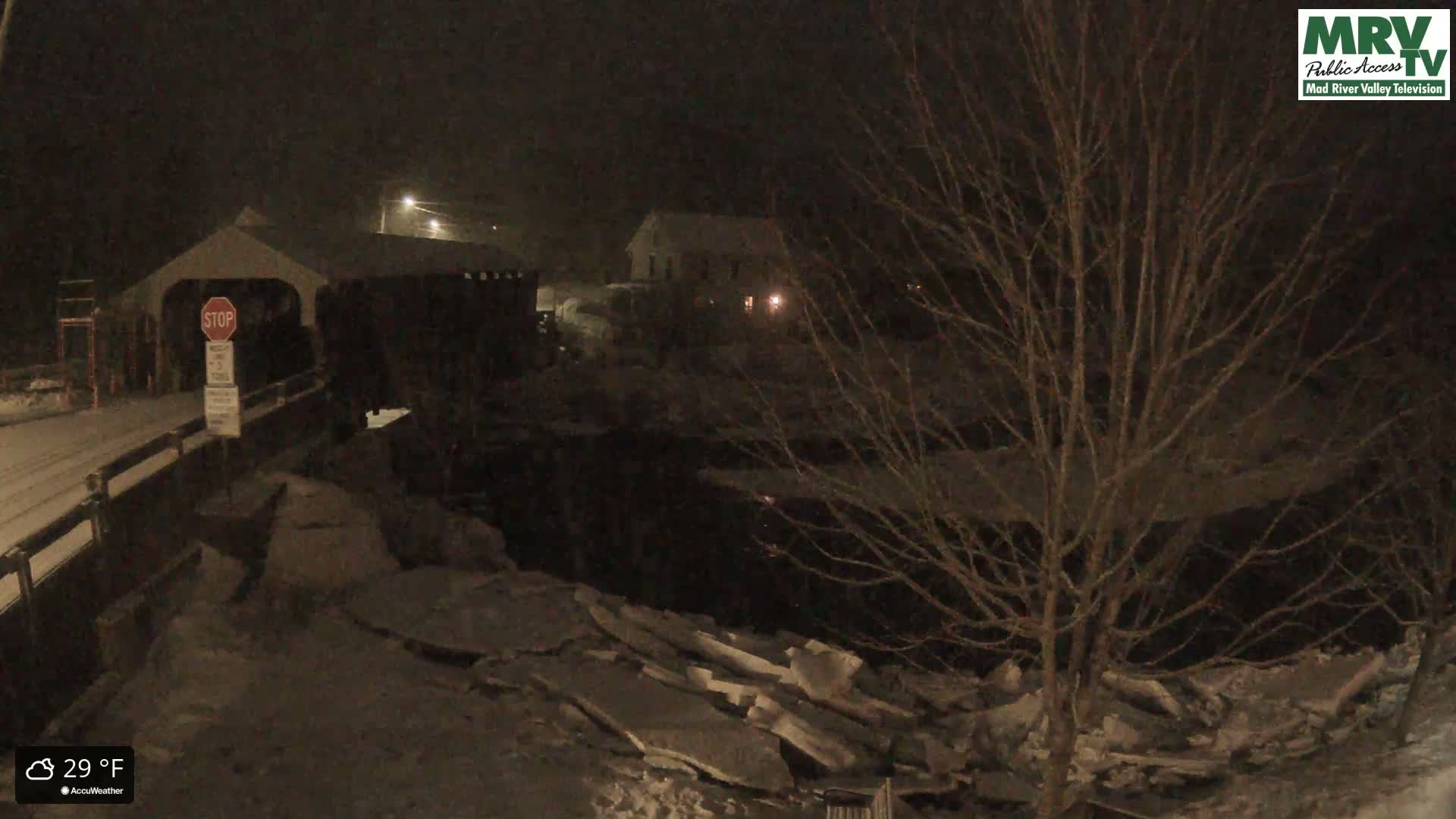 A snow-covered covered bridge and nearby building are visible at night under falling snow.
