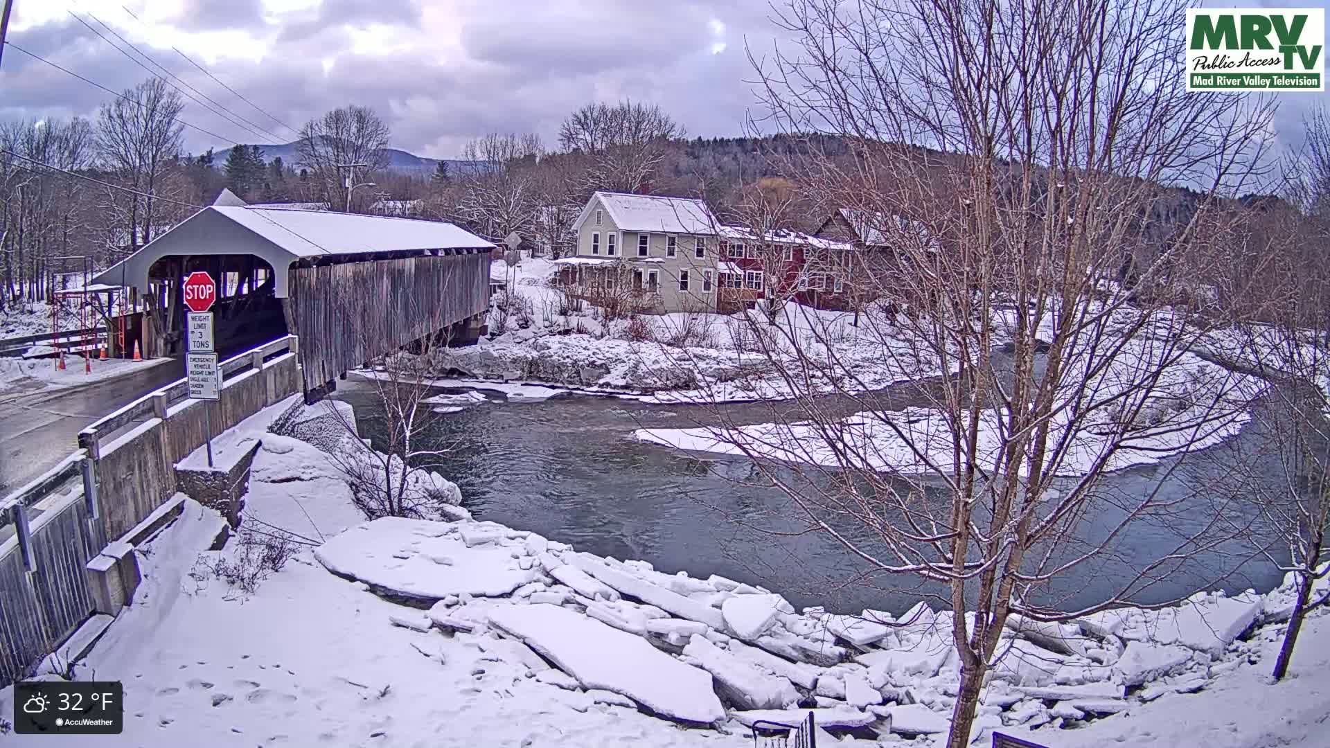 A snow-covered wooden covered bridge spans a partially frozen river in a winter scene, with houses and bare trees visible in the background under a cloudy sky.