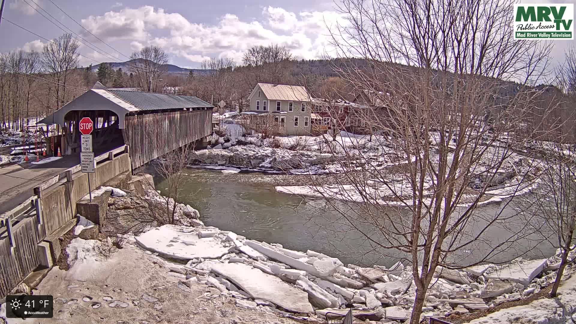 A covered bridge spans a river partially covered with broken ice floes under a partly sunny sky.