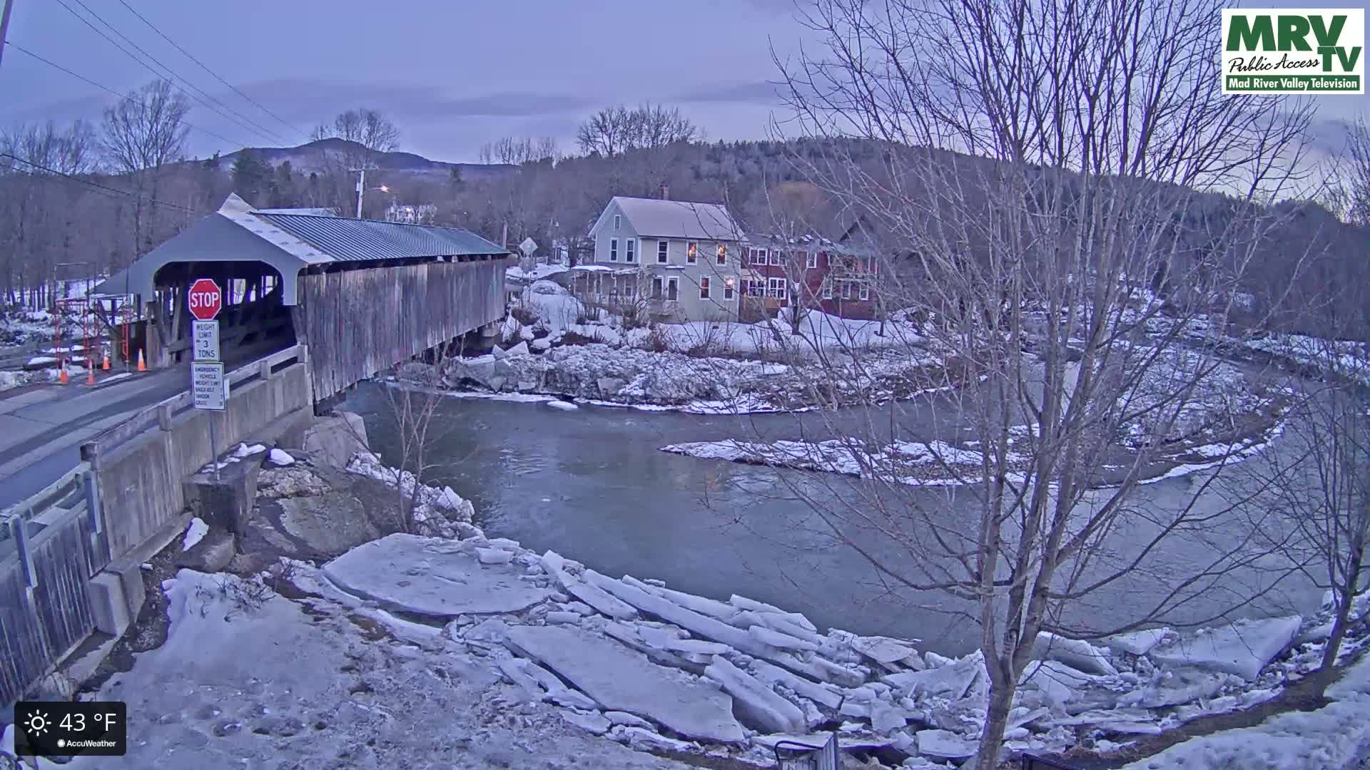 A covered bridge spans a partially ice-covered river, with snow-covered banks and houses visible in the background under a twilight sky.