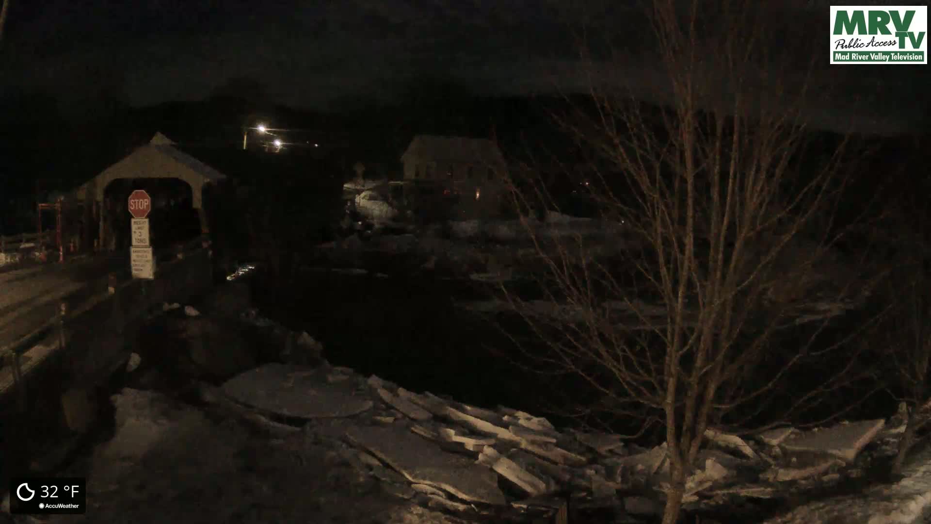 A nighttime view of a covered bridge with ice floes in a river below, under a dark, clear sky at 32 degrees Fahrenheit.