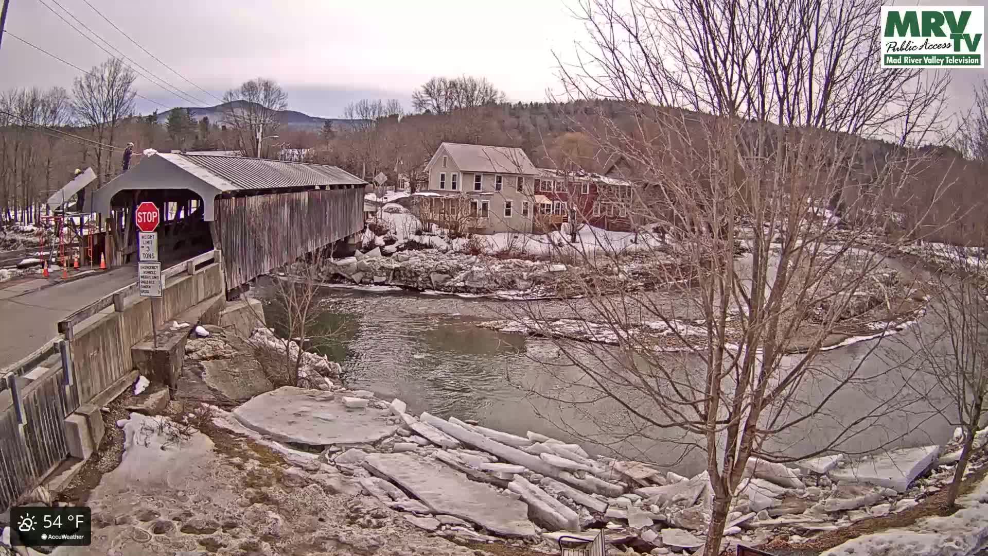 A covered bridge stands over a river with partially melted ice along the banks, under a cloudy sky.