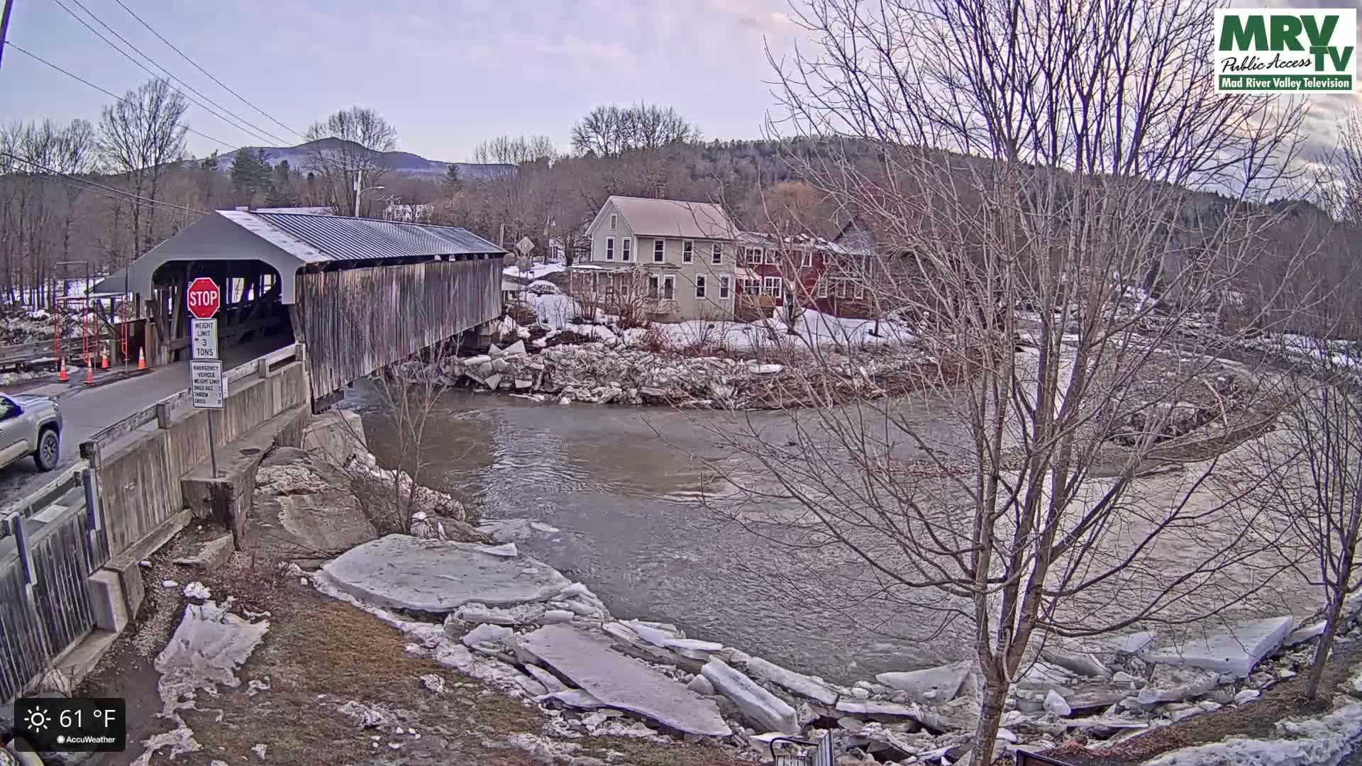 A covered bridge spans a river with broken ice floes along its banks, under a mostly clear sky, near some buildings on a partly snowy, winter day.