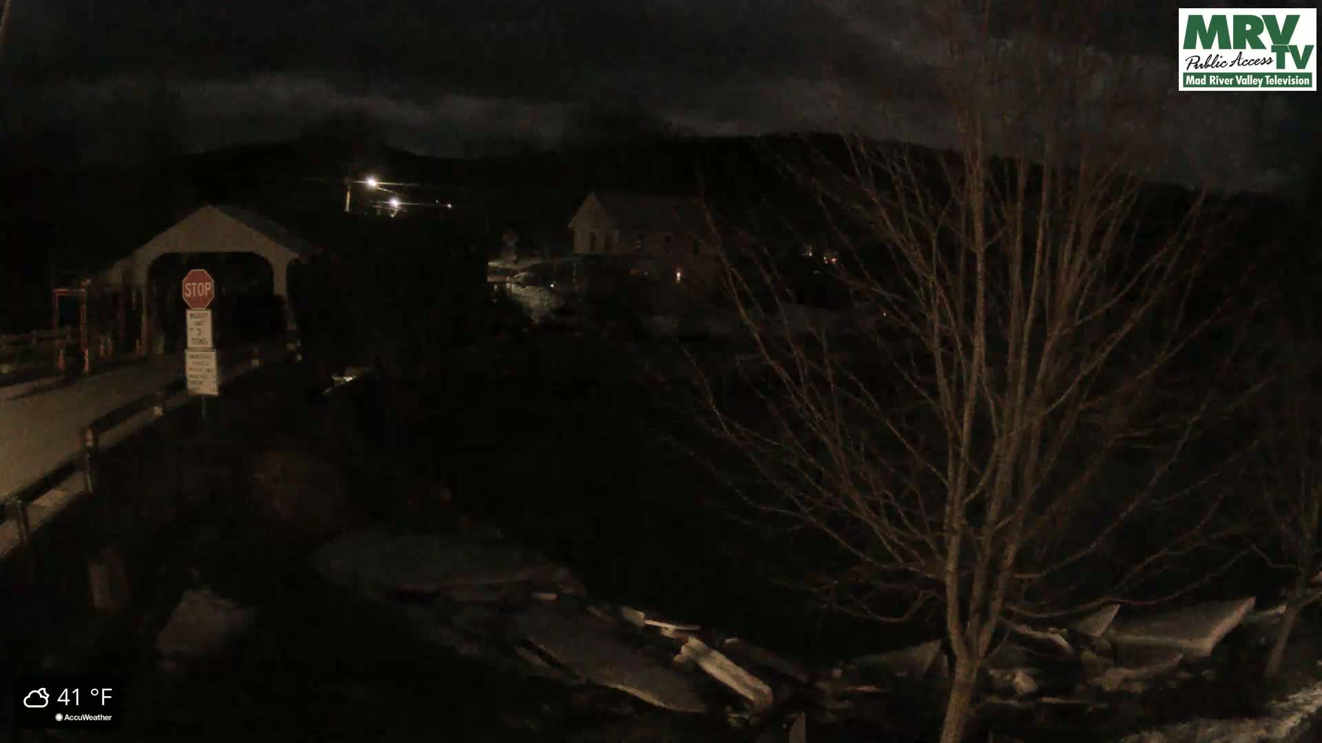 A dark nighttime scene shows a covered bridge, a building in the distance, and bare trees under a cloudy sky, with the temperature at 41°F.