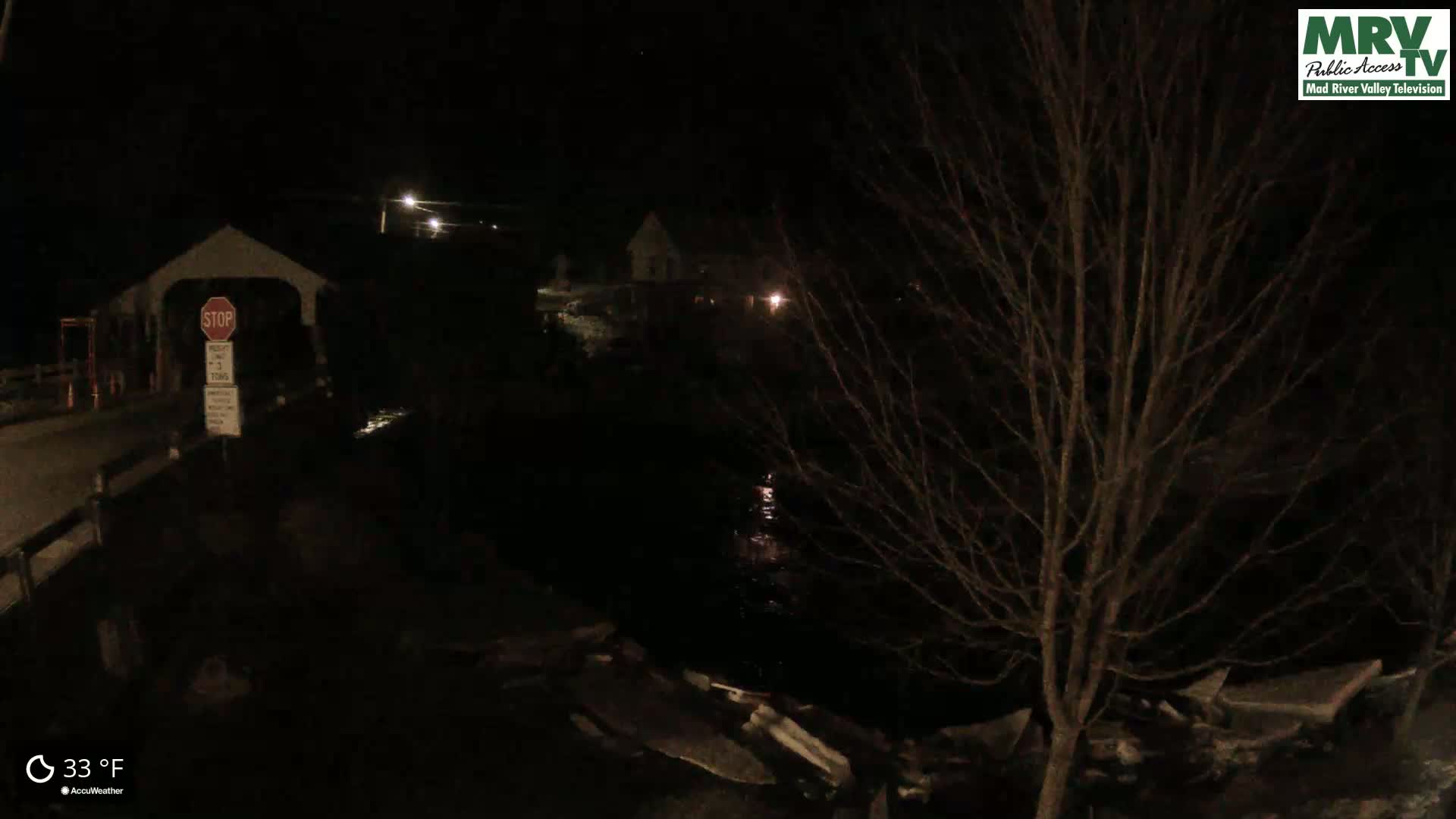 A nighttime view of a covered bridge, a dark river, and bare trees at 33°F.