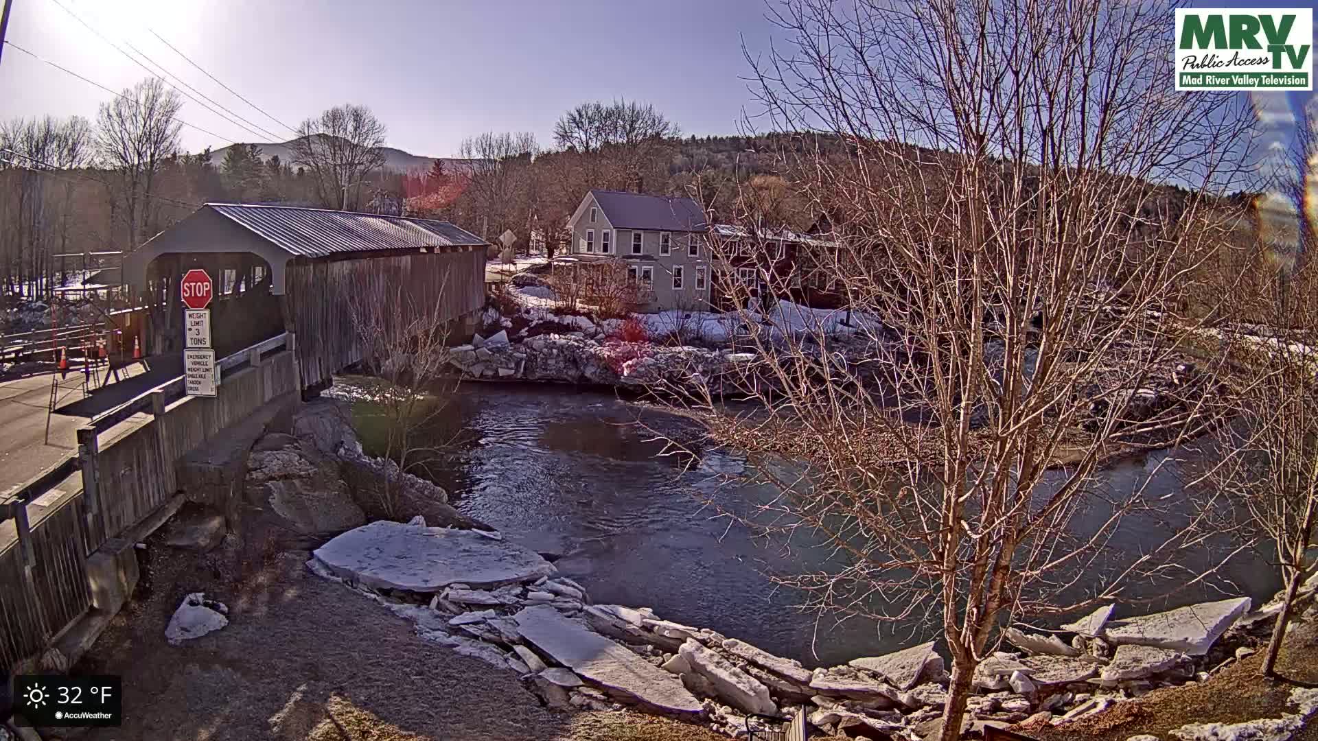 A wooden covered bridge spans a river with partially melted ice flows along its banks on a sunny day.