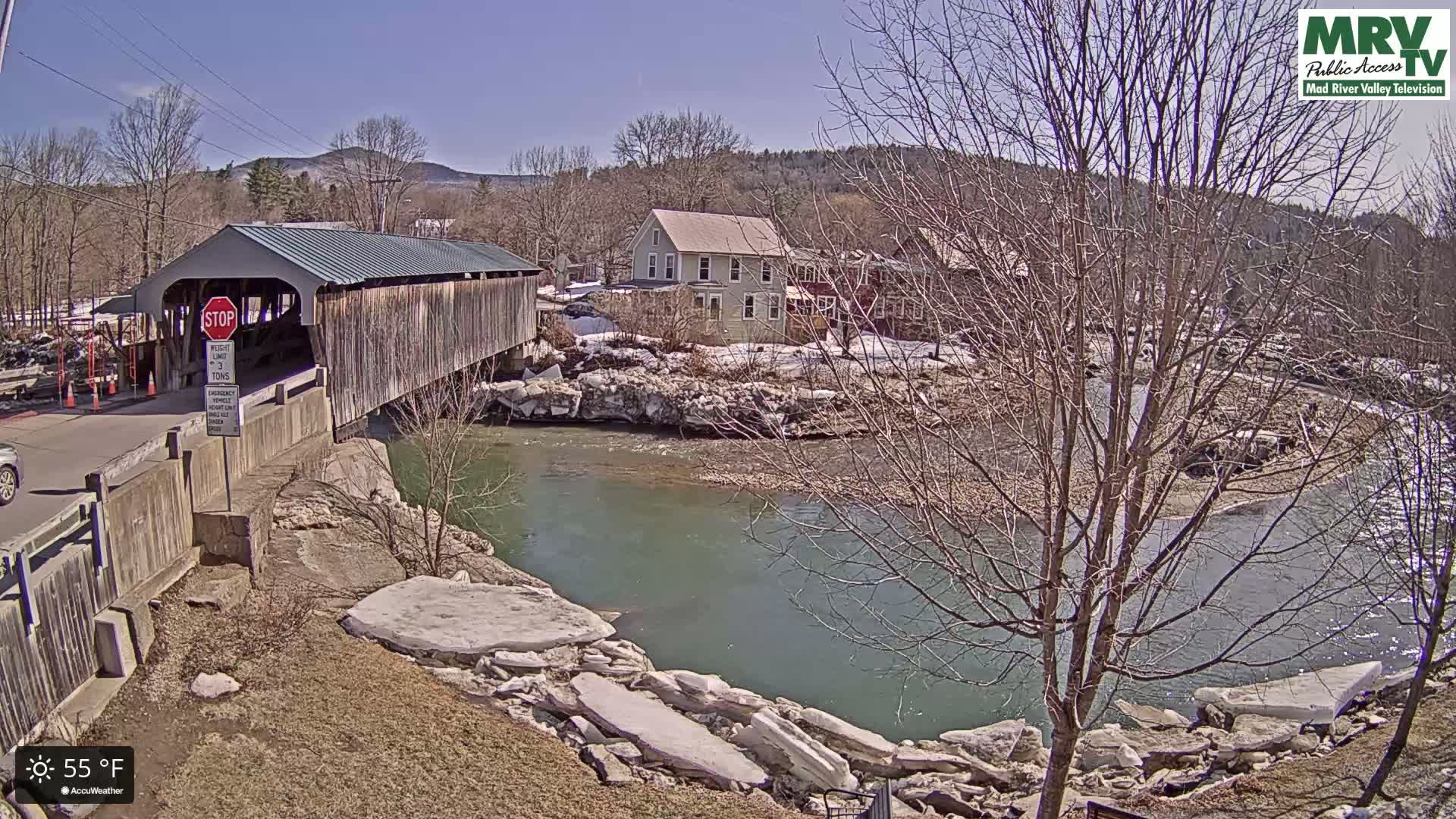 A covered bridge spans a river with partially melted ice along its banks under a sunny sky.