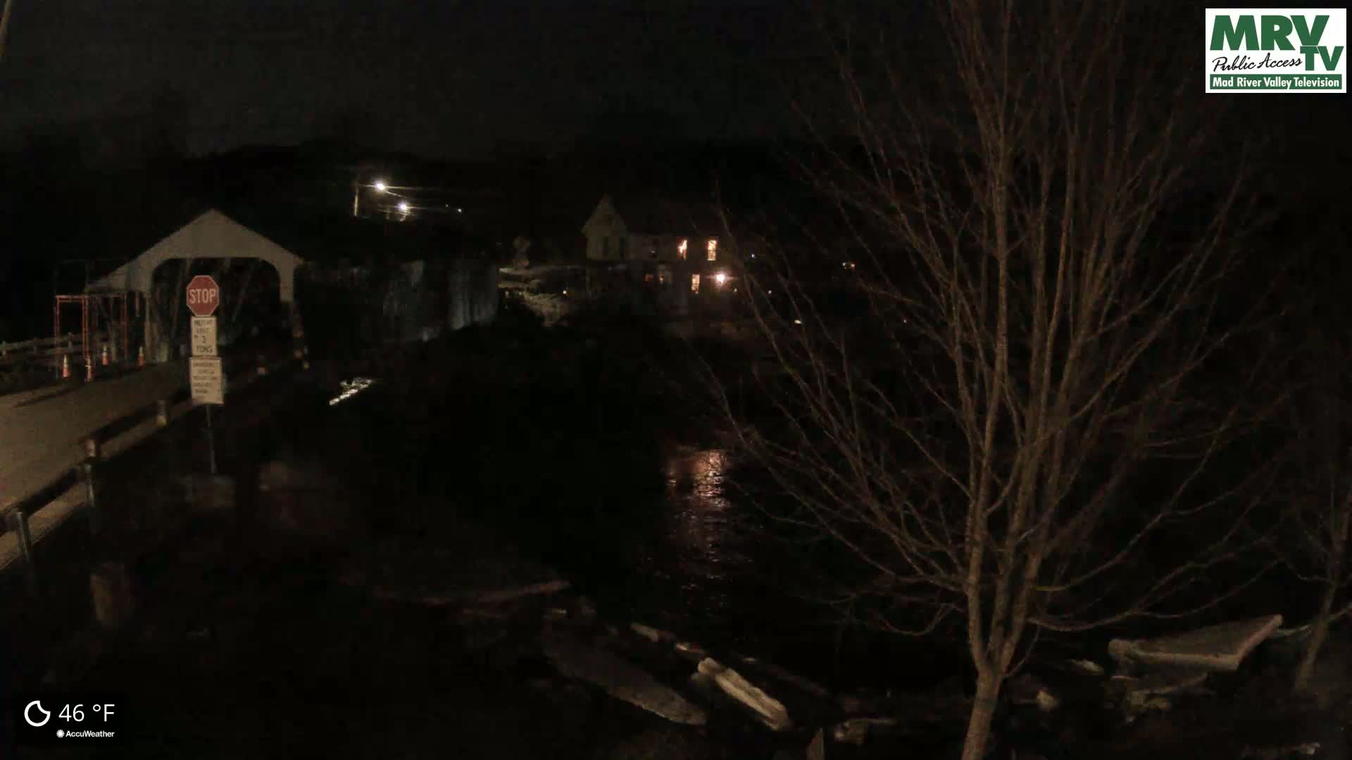 A nighttime view of a covered bridge under construction, a house with lit windows, and a dark river, with the temperature at 46°F.
