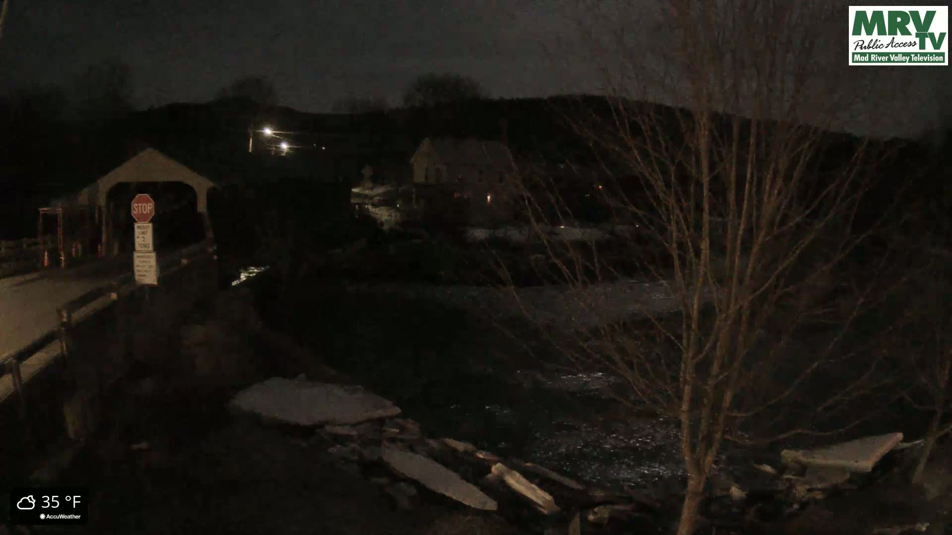 A nighttime view of a covered bridge, a river with some ice, and a building beyond the river, under a dark sky with a temperature of 35°F.