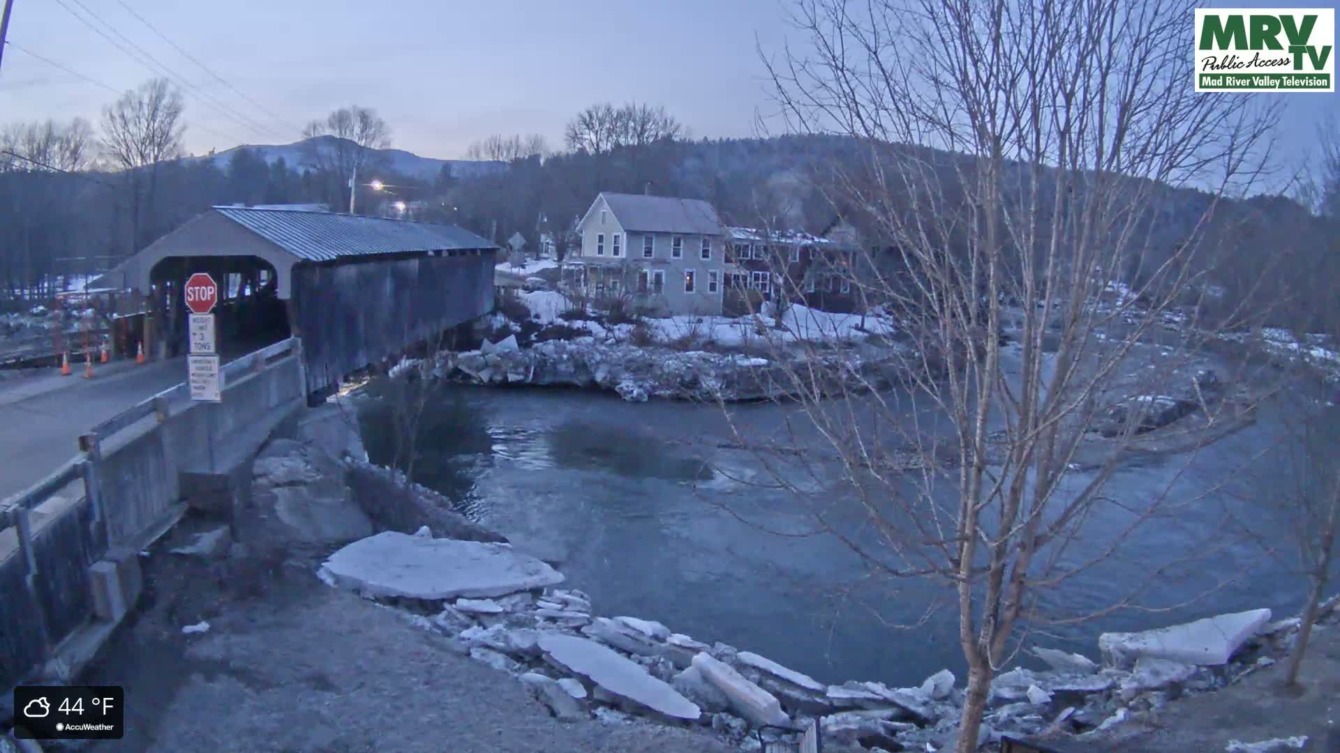 A covered bridge spans a river partially covered in ice under a twilight sky.
