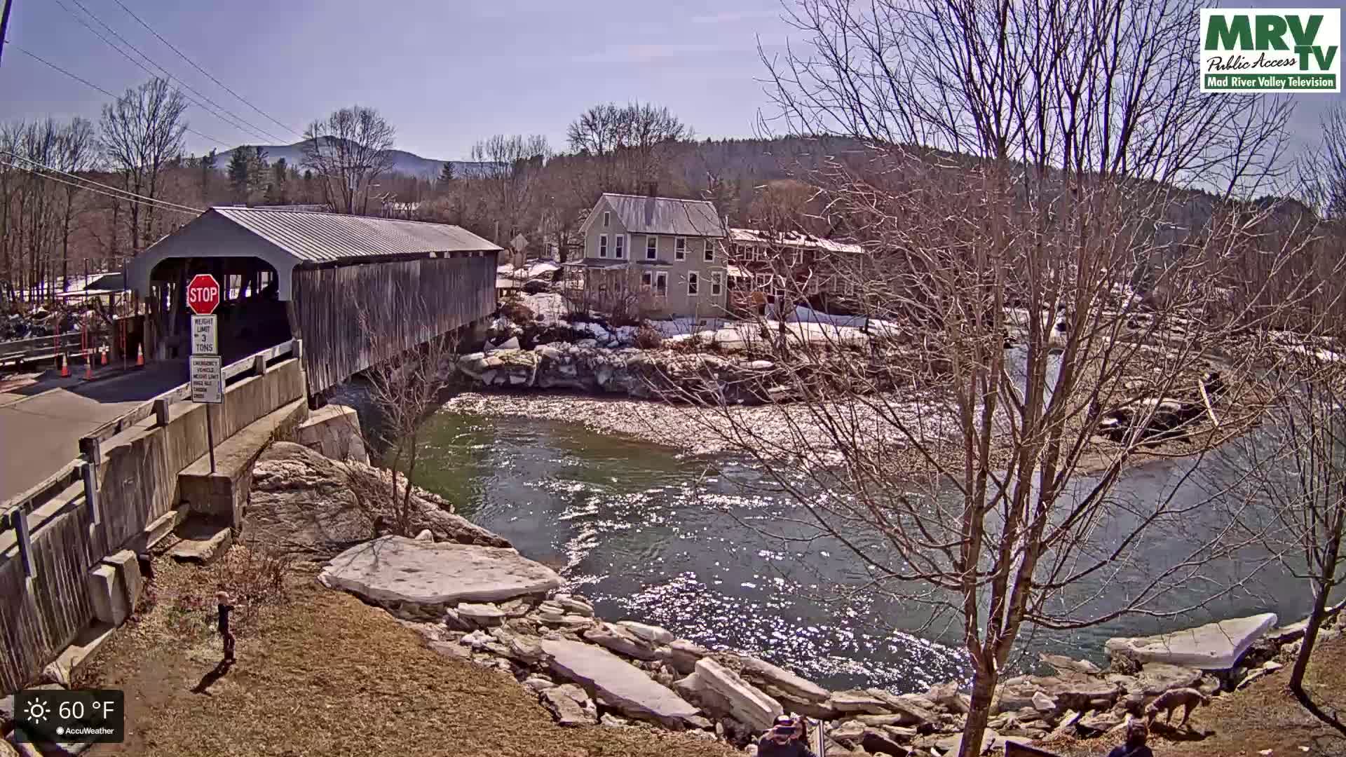 A covered bridge spans a flowing river on a sunny 60°F day, with a house and some remaining snow visible on the riverbank.