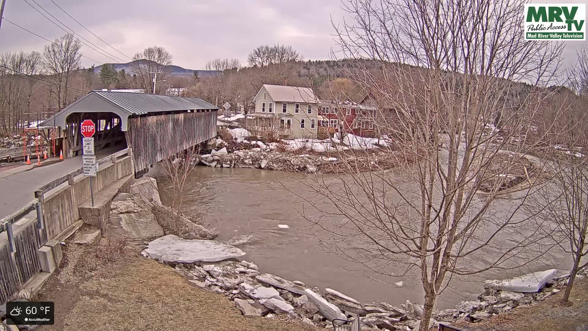 A covered bridge stands over a muddy, flowing river partially covered in ice, with buildings and bare trees lining the banks under an overcast sky.
