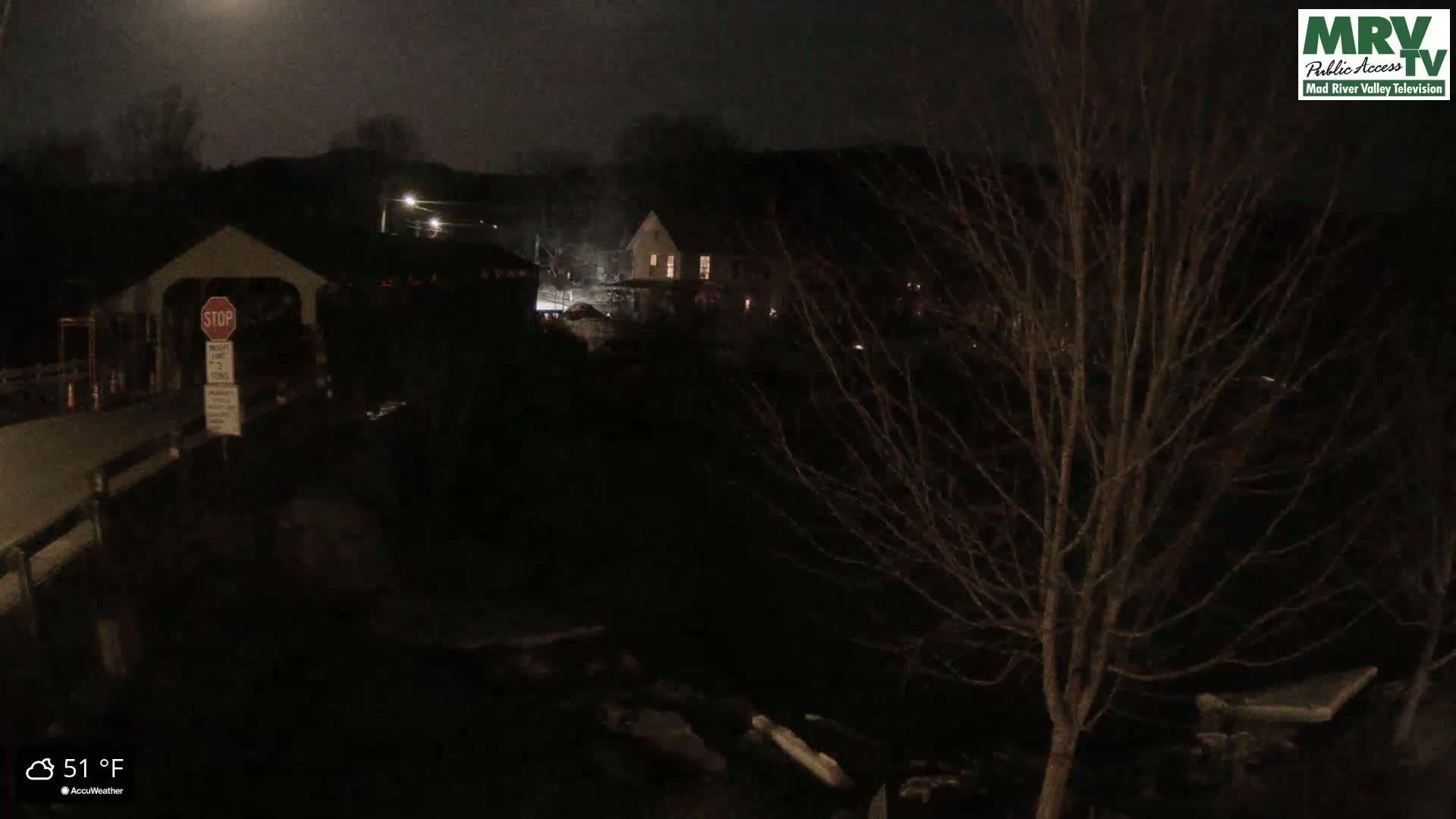 A nighttime view of a covered bridge and a house, under a dark, cloudy sky, with a temperature of 51°F.