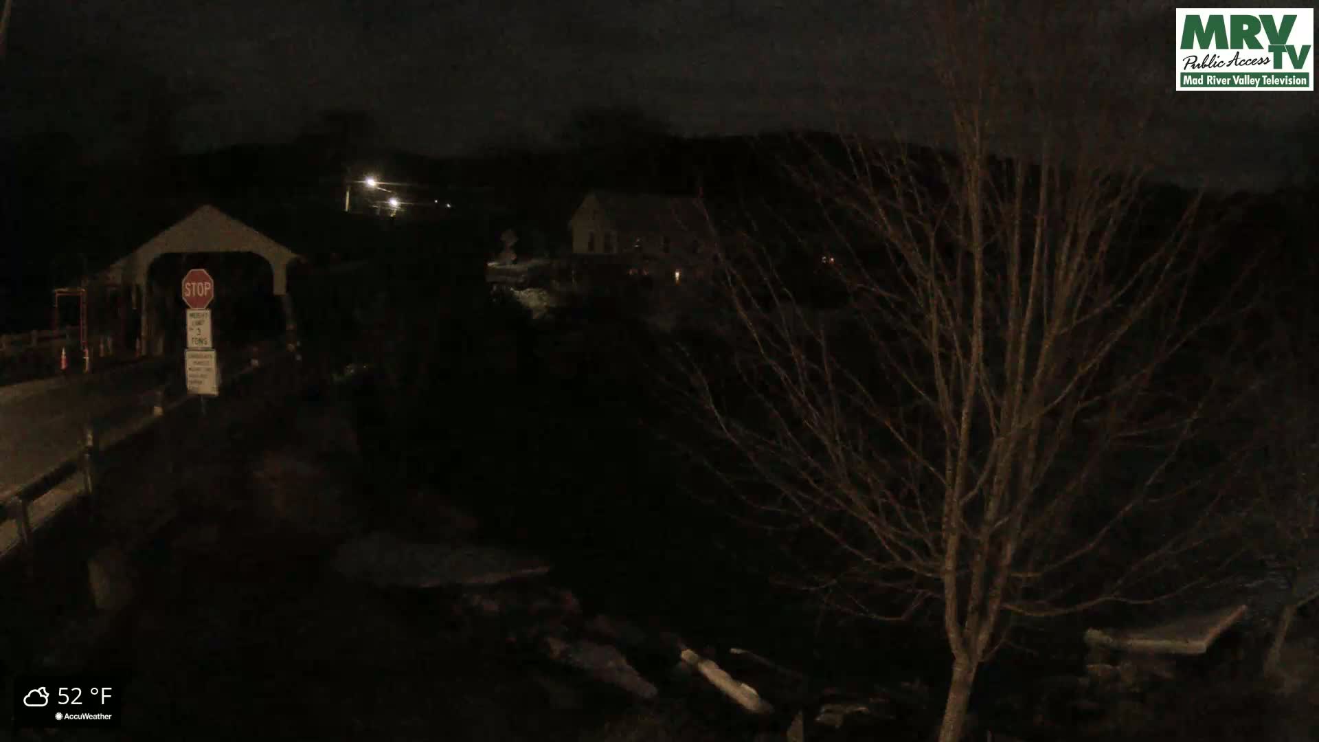 A nighttime view of a covered bridge and a building, with a bare tree in the foreground and a temperature of 52 degrees Fahrenheit.