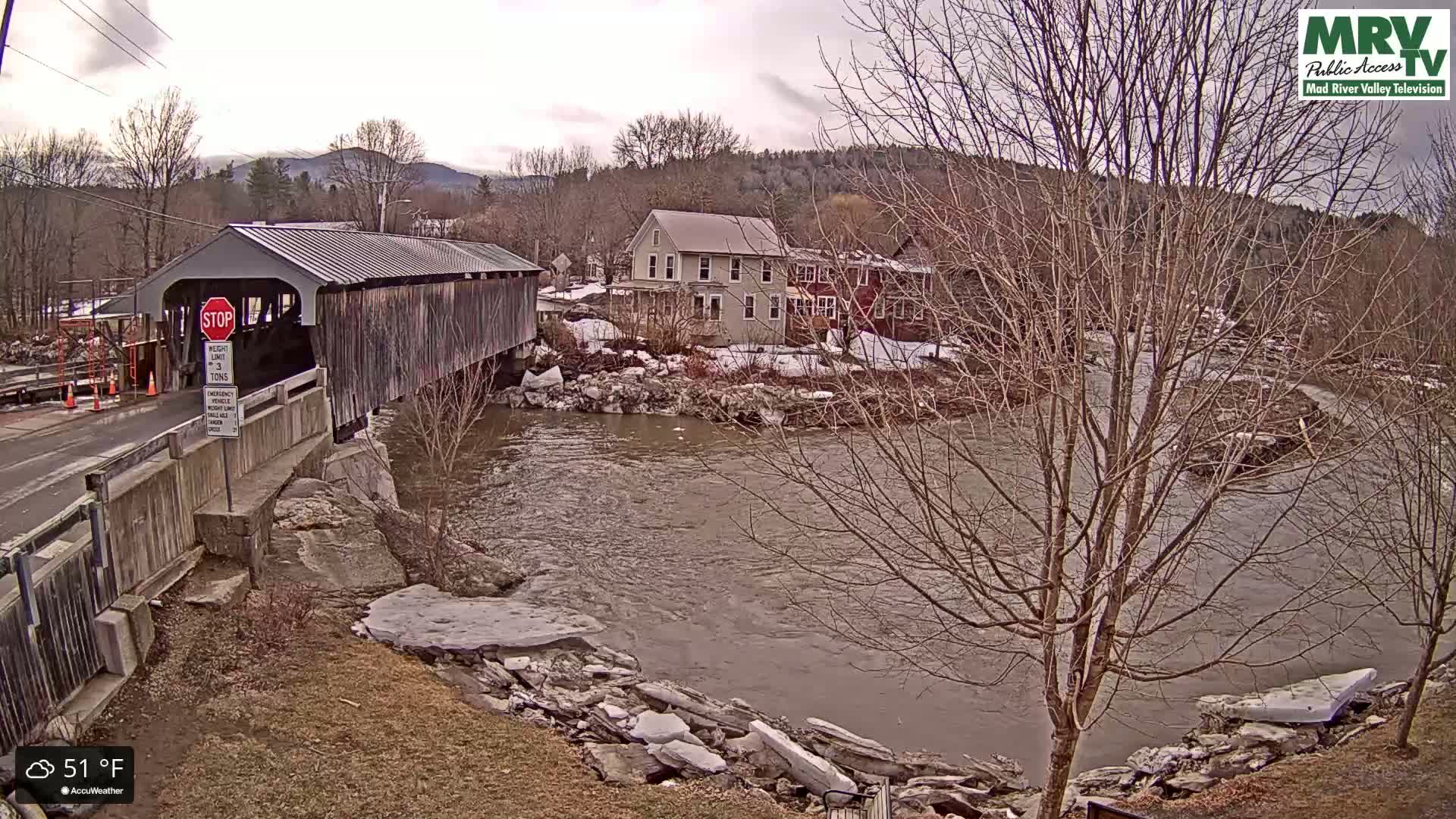 A covered bridge spans a brown river, with buildings and bare trees lining the banks on an overcast day at 51°F.