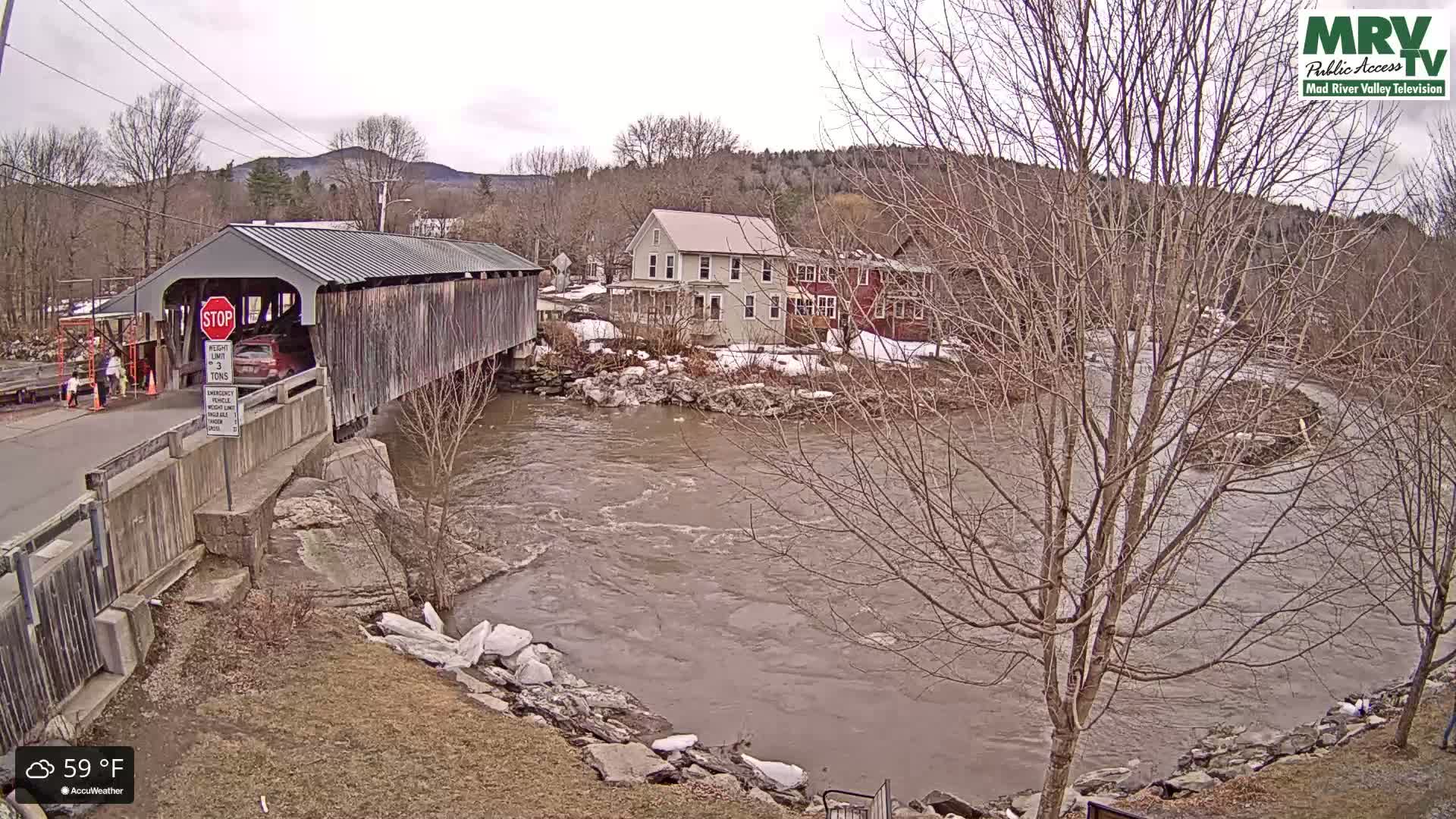 A covered bridge spans a muddy, swiftly flowing river under an overcast sky, with bare trees and buildings lining the riverbank.