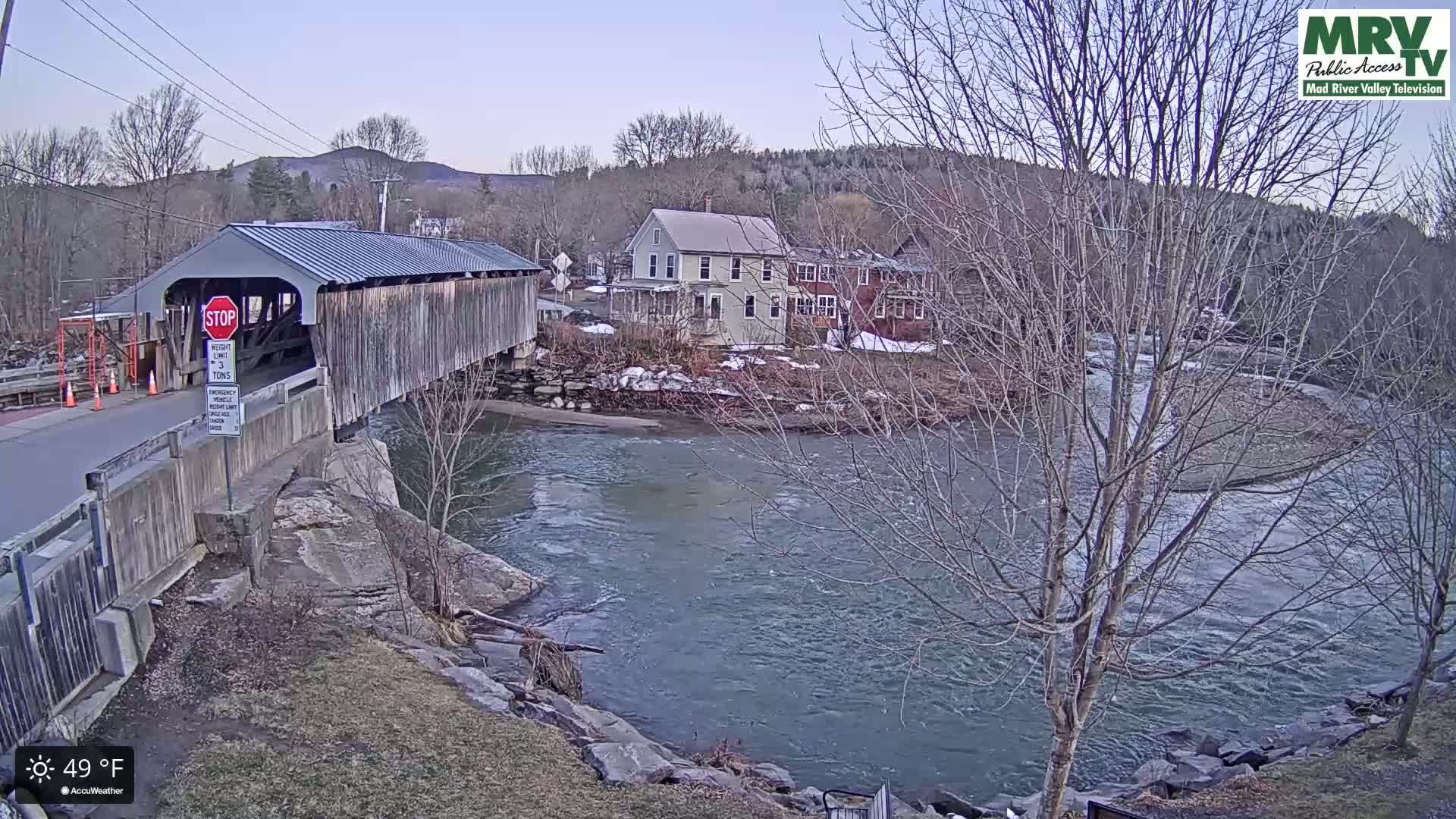 A covered bridge spans a river, with houses and bare trees visible on the far bank under a mostly clear sky.