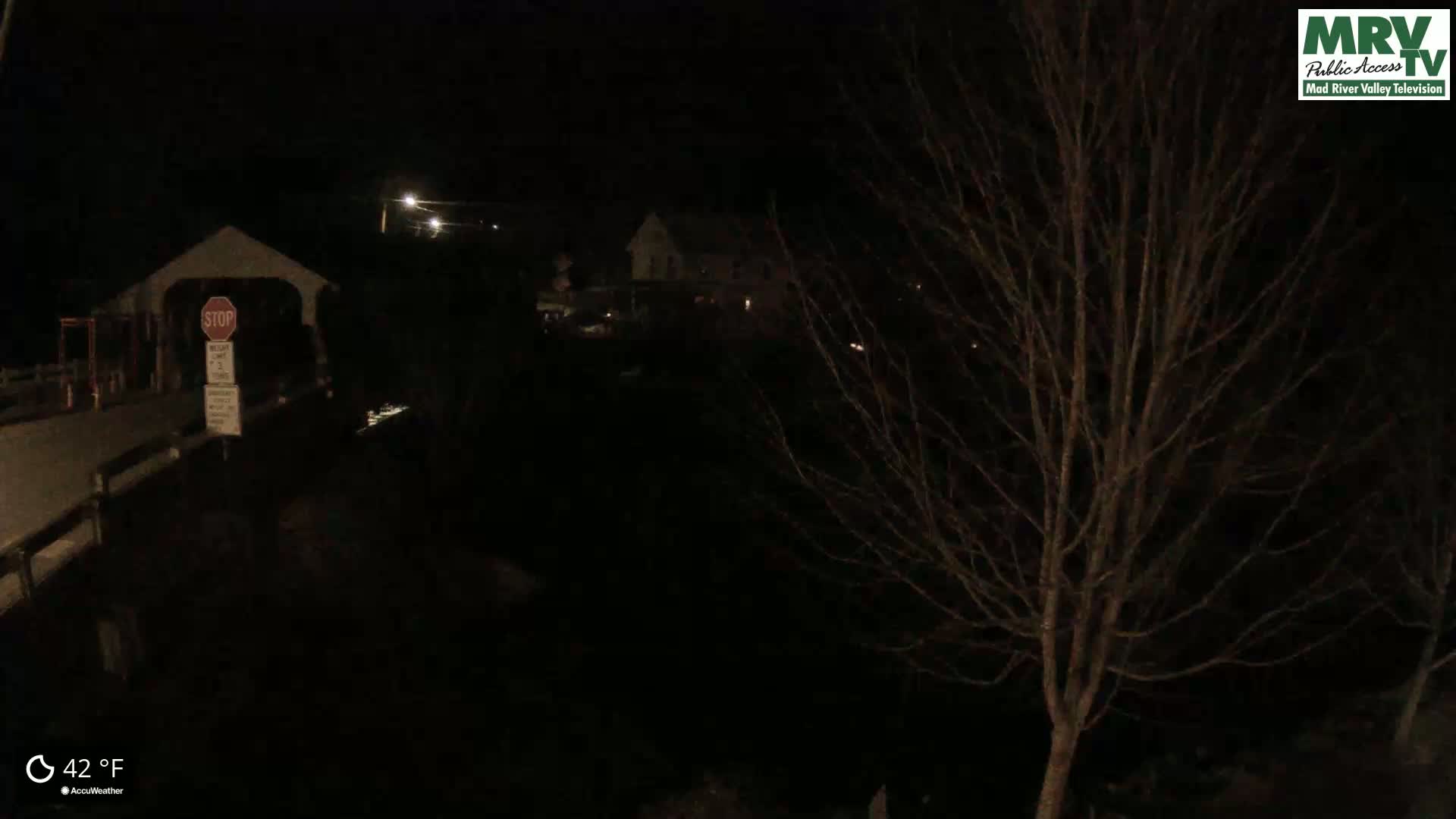 A nighttime view of a covered bridge and a building under a dark sky, with a bare tree in the foreground and a temperature of 42°F.