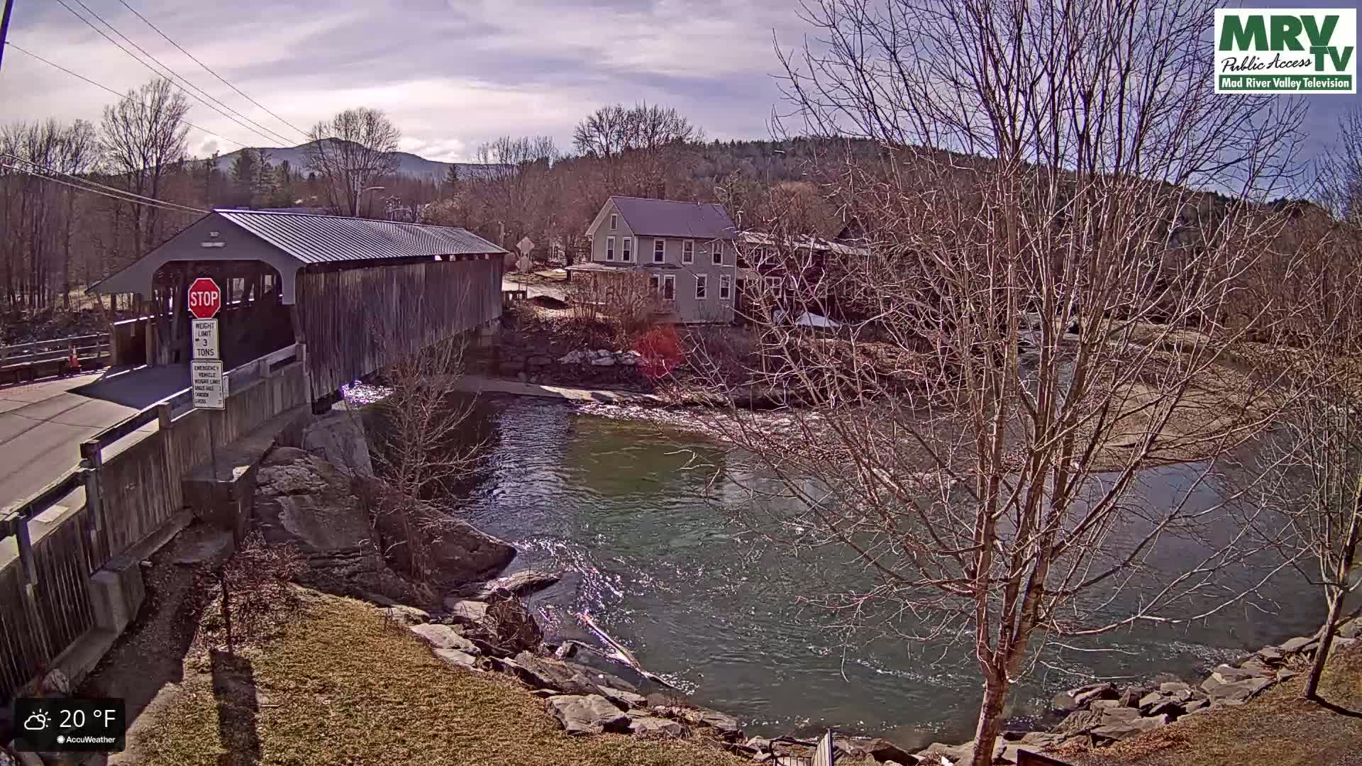 A covered bridge spans a river on a partly sunny, 20°F day, with bare trees lining the riverbanks and a building visible in the background.