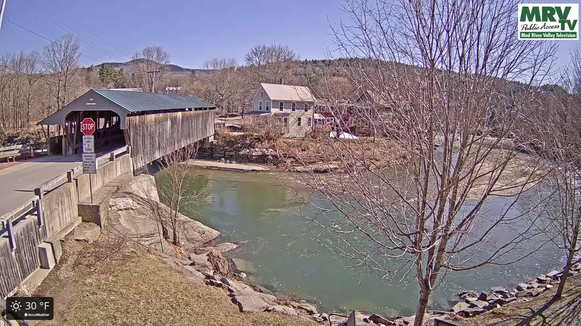 A covered bridge spans a river under a clear, sunny sky, with leafless trees lining the banks and a small town visible in the background.
