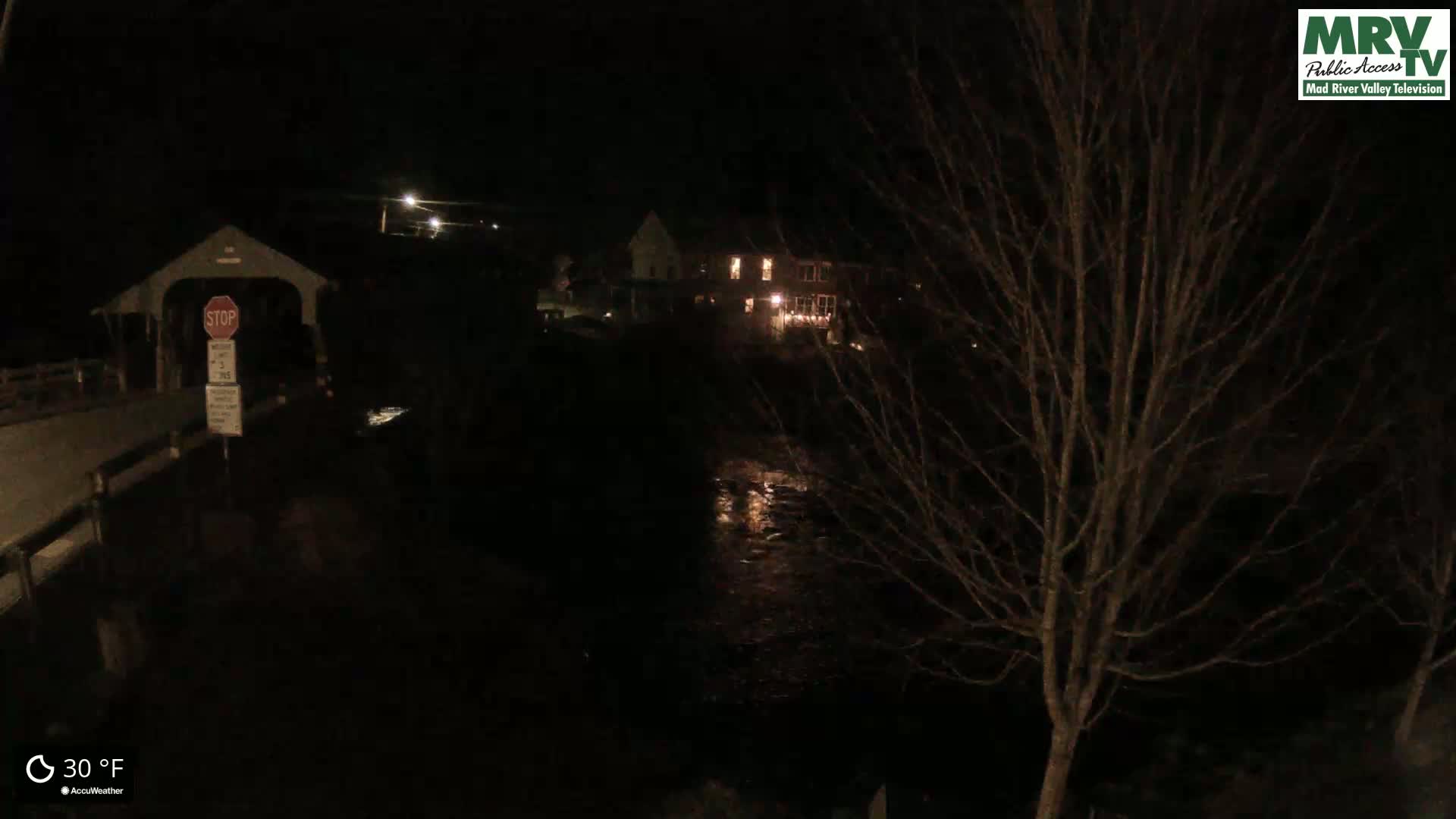 A nighttime view of a covered bridge, a calm river, and a sparsely lit building, under a dark, clear sky with a temperature of 30°F.