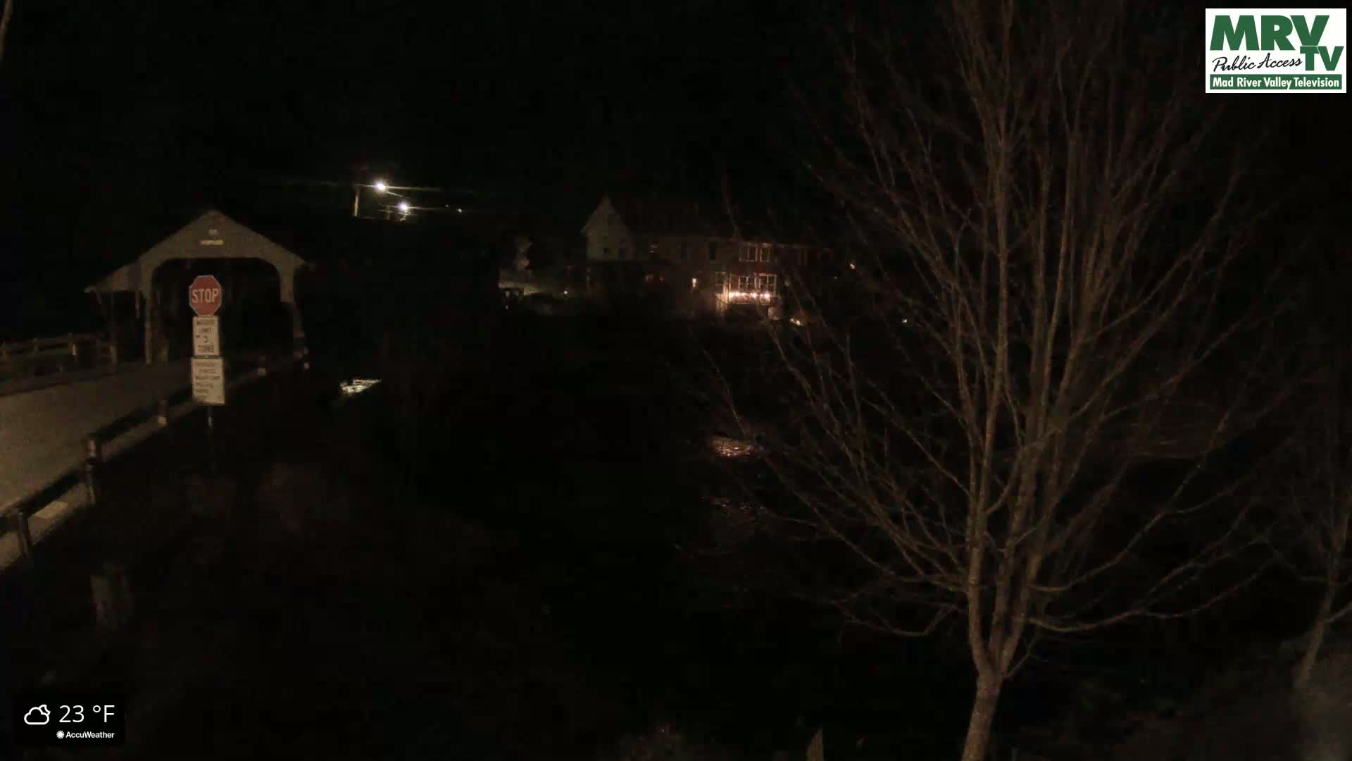 A nighttime view of a covered bridge and buildings across a dark body of water, under a clear, 23-degree Fahrenheit sky.