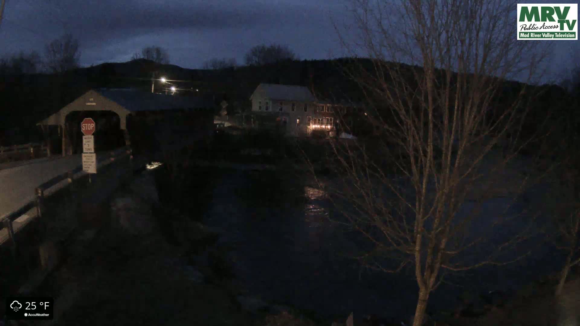 A dark, nighttime scene shows a covered bridge over a river, with a building and trees visible in the background under a mostly clear, dark sky at 25°F.
