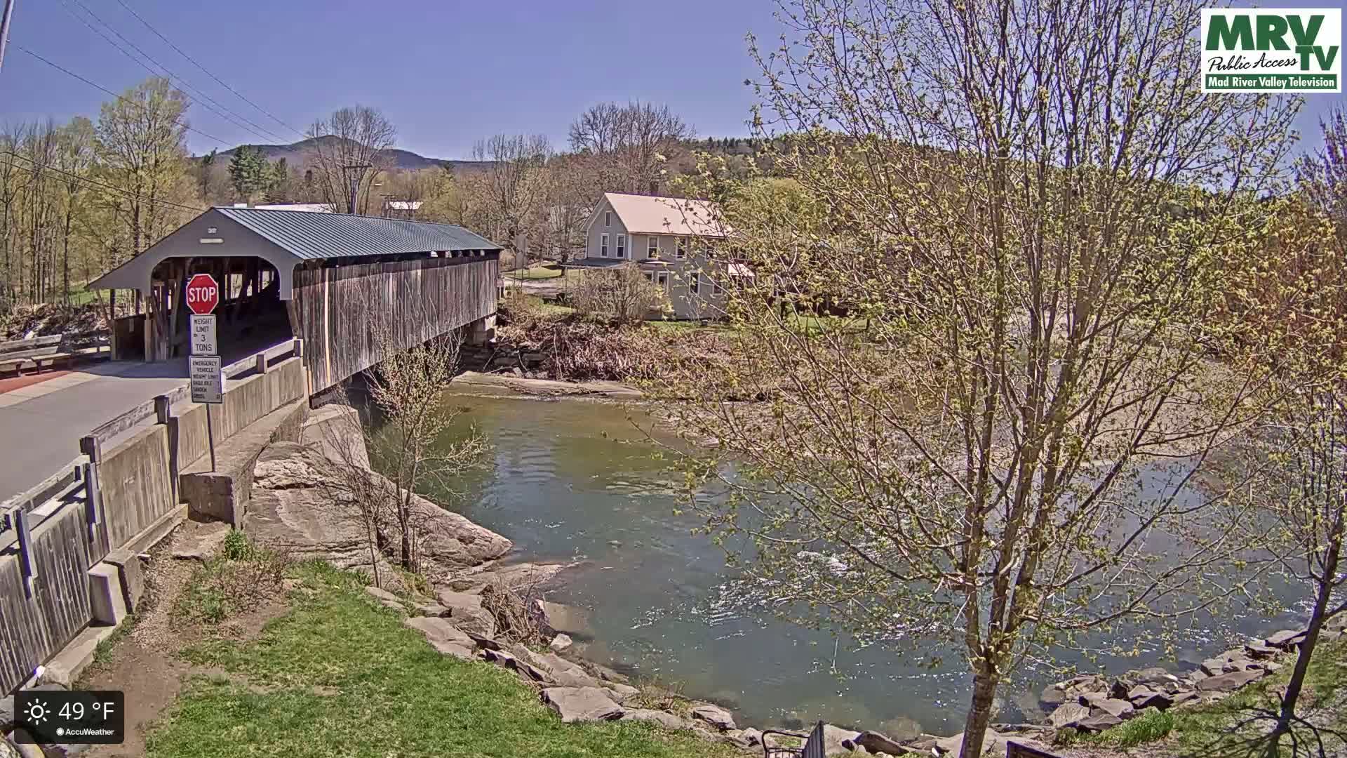 A wooden covered bridge spans a river on a sunny day, with budding trees and a house visible in the background.