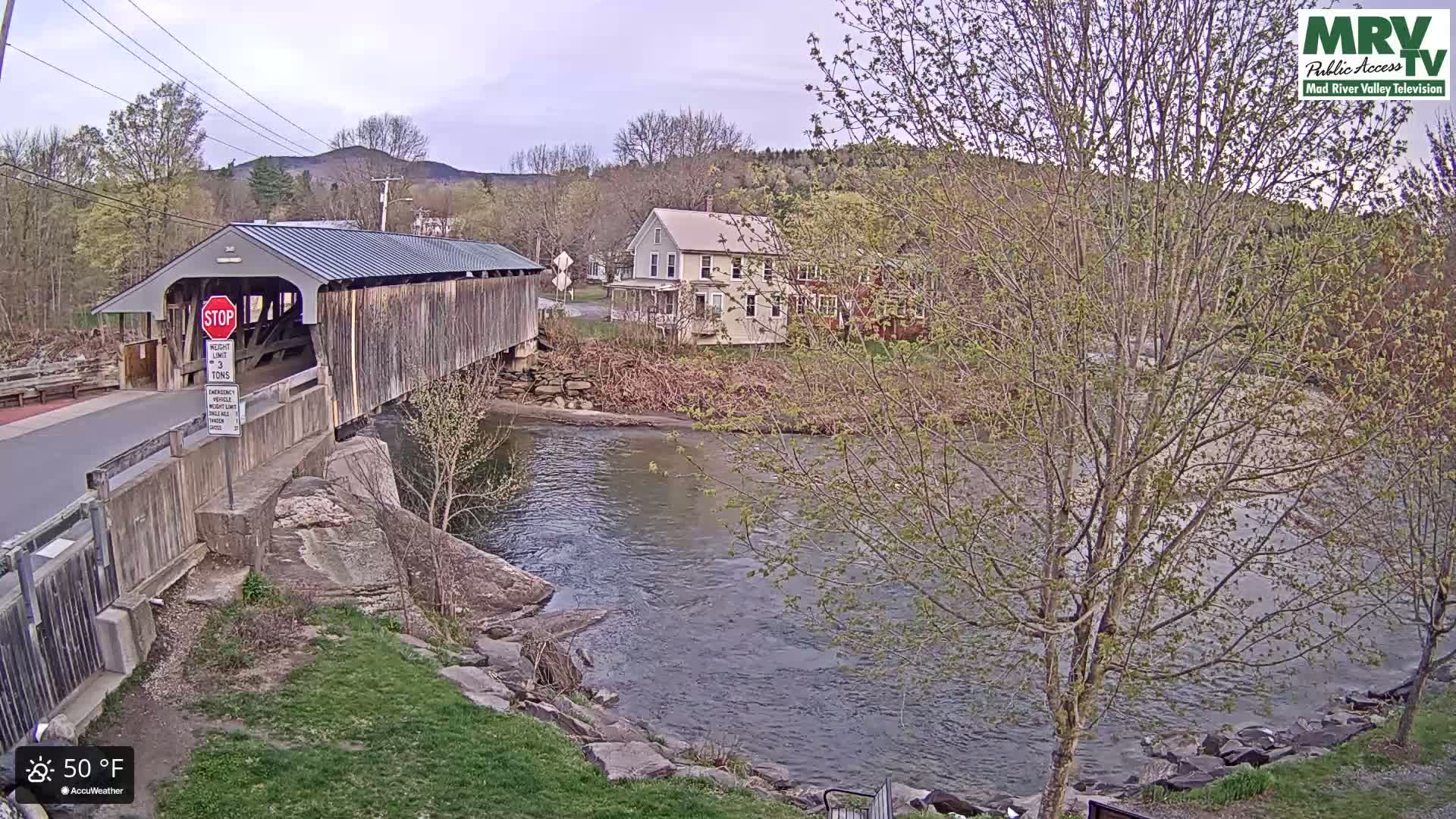 A covered bridge spans a river under a partly cloudy sky, with houses and trees visible on the banks, and the temperature is 50°F.