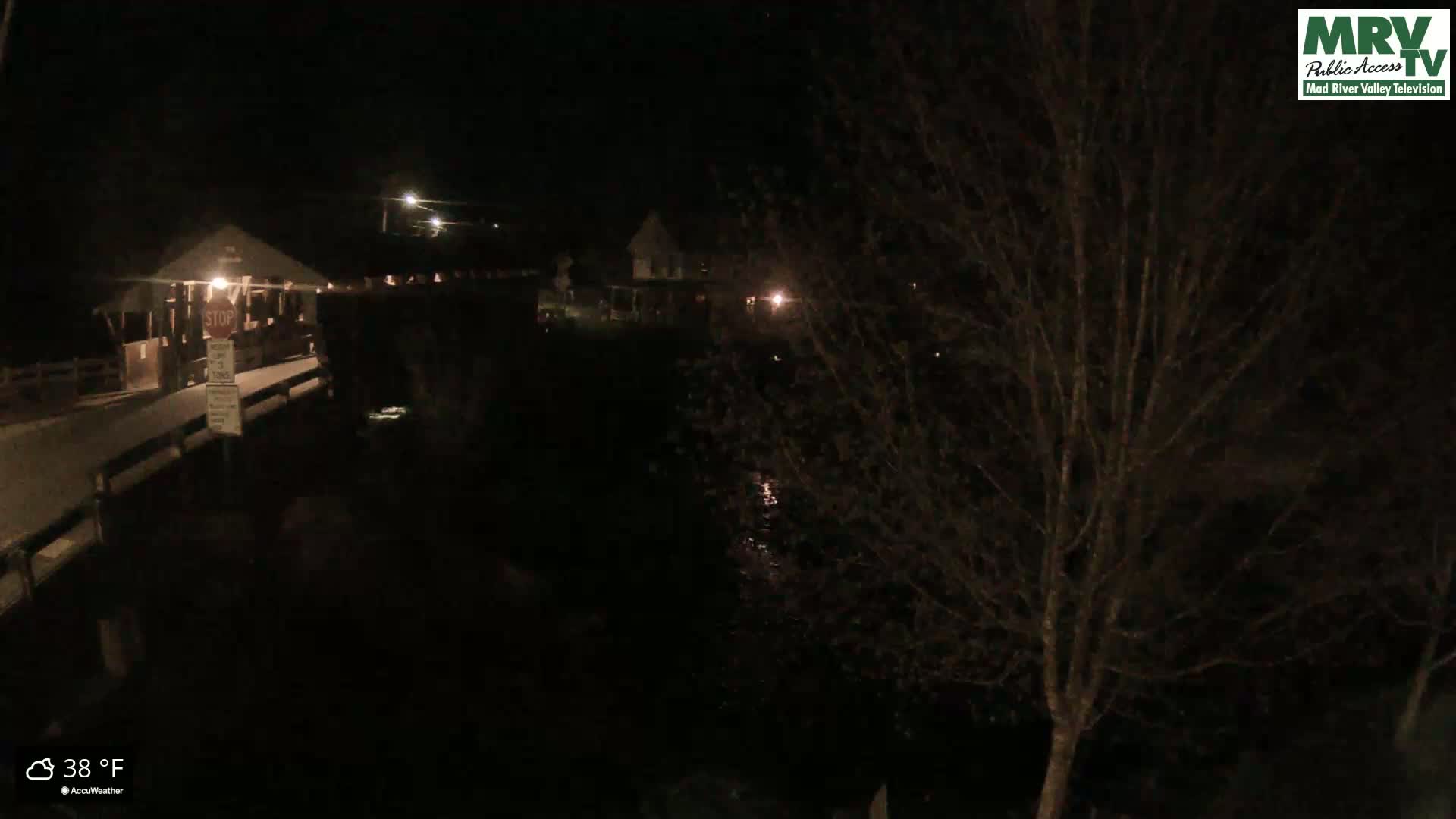 A nighttime view of a covered bridge and nearby buildings, with a tree in the foreground, at 38°F.