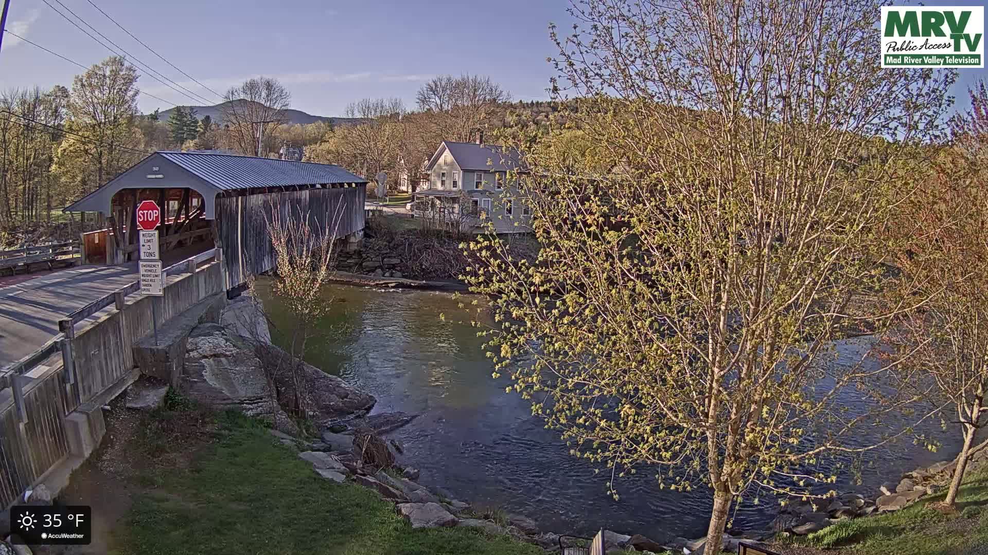 A covered bridge spans a calm river under a clear, sunny sky, with budding trees and a house visible in the background.