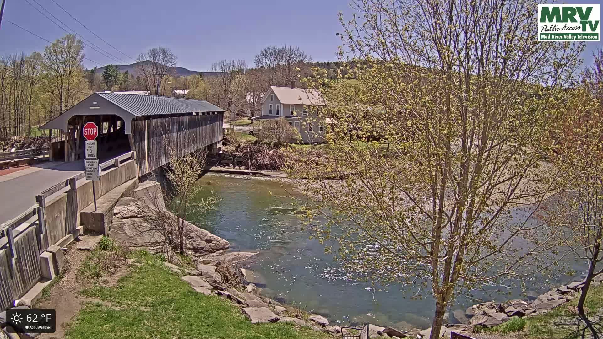 A wooden covered bridge spans a river under a sunny sky, with budding trees and a house visible in the background.