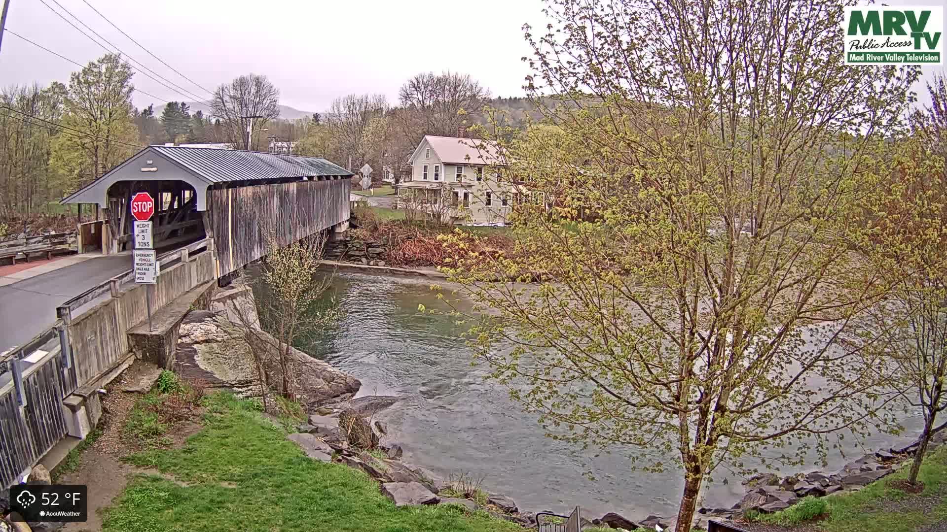 A covered bridge spans a river under an overcast sky, with budding trees and a house visible in the background.