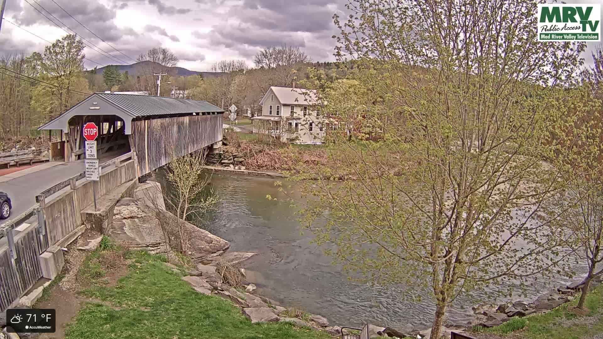 A covered bridge spans a calm river under a partly cloudy sky, with a house and trees visible in the background.