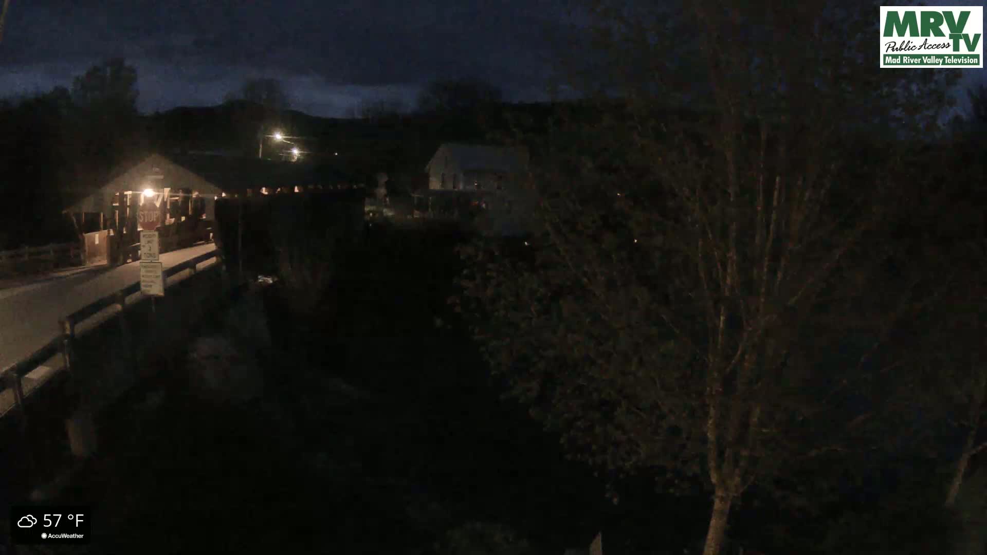 A dark, nighttime view of a covered bridge and a building, under a mostly cloudy sky.