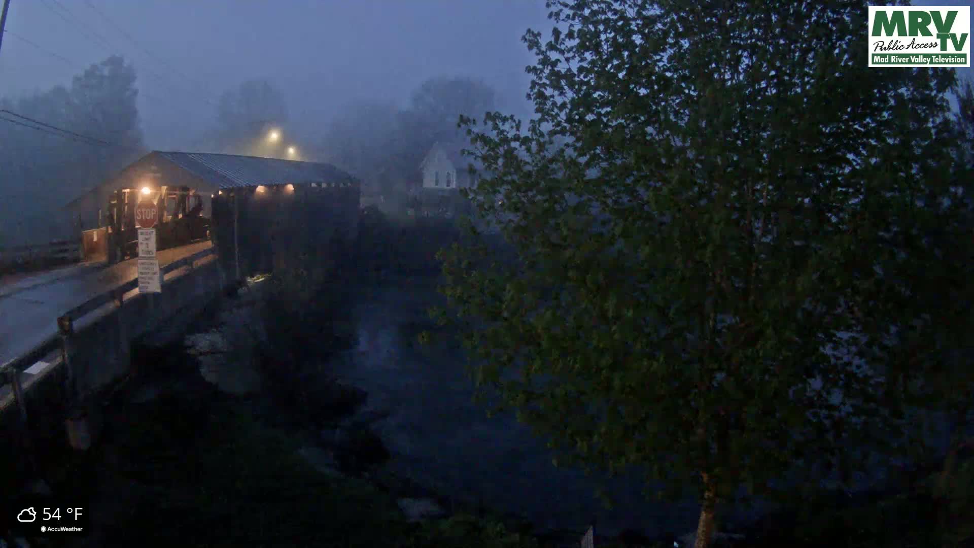 A covered bridge is dimly lit in the fog at night, near a body of water and trees.