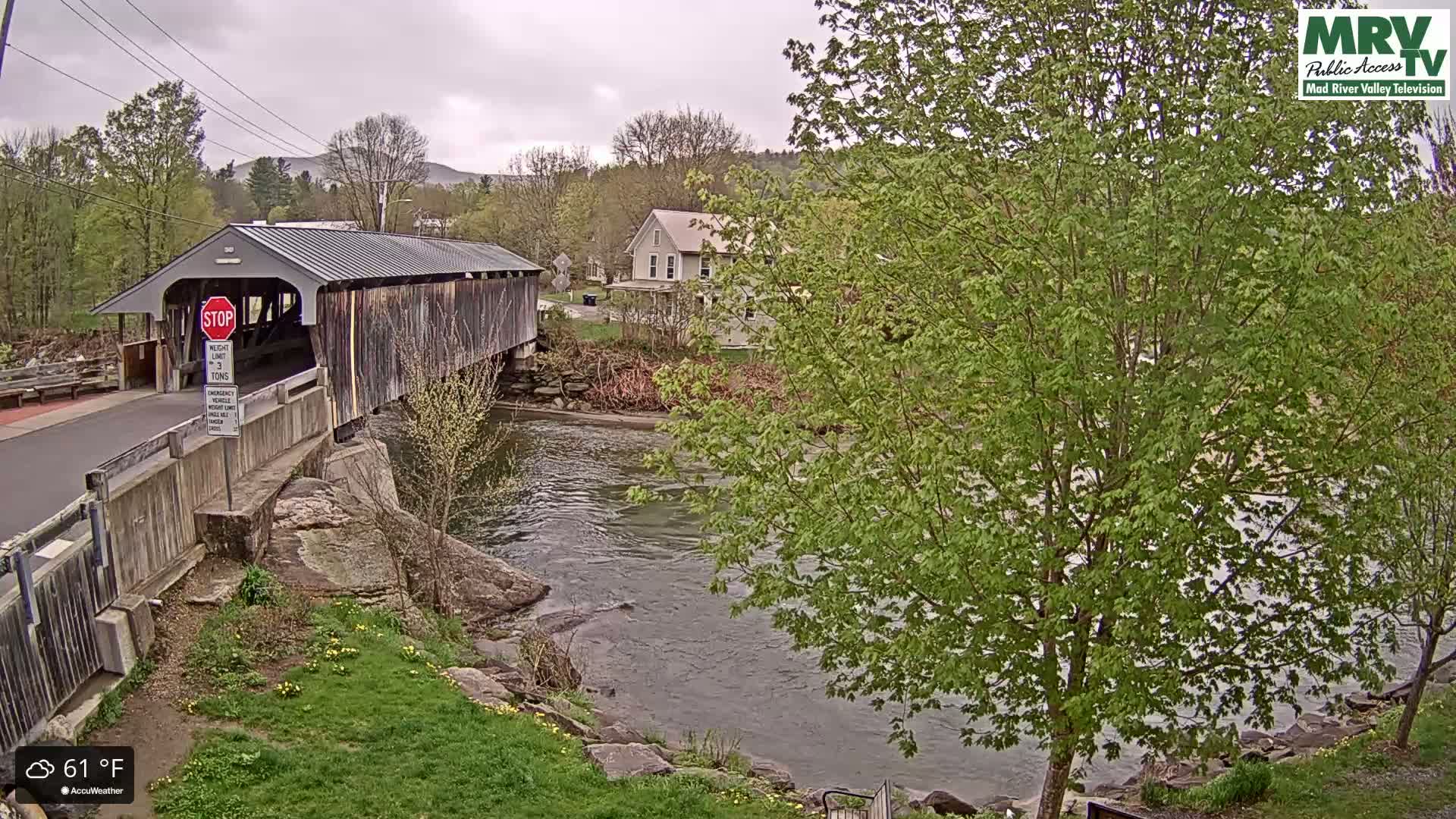 A covered bridge spans a river under an overcast sky, with green trees and a house visible in the background.