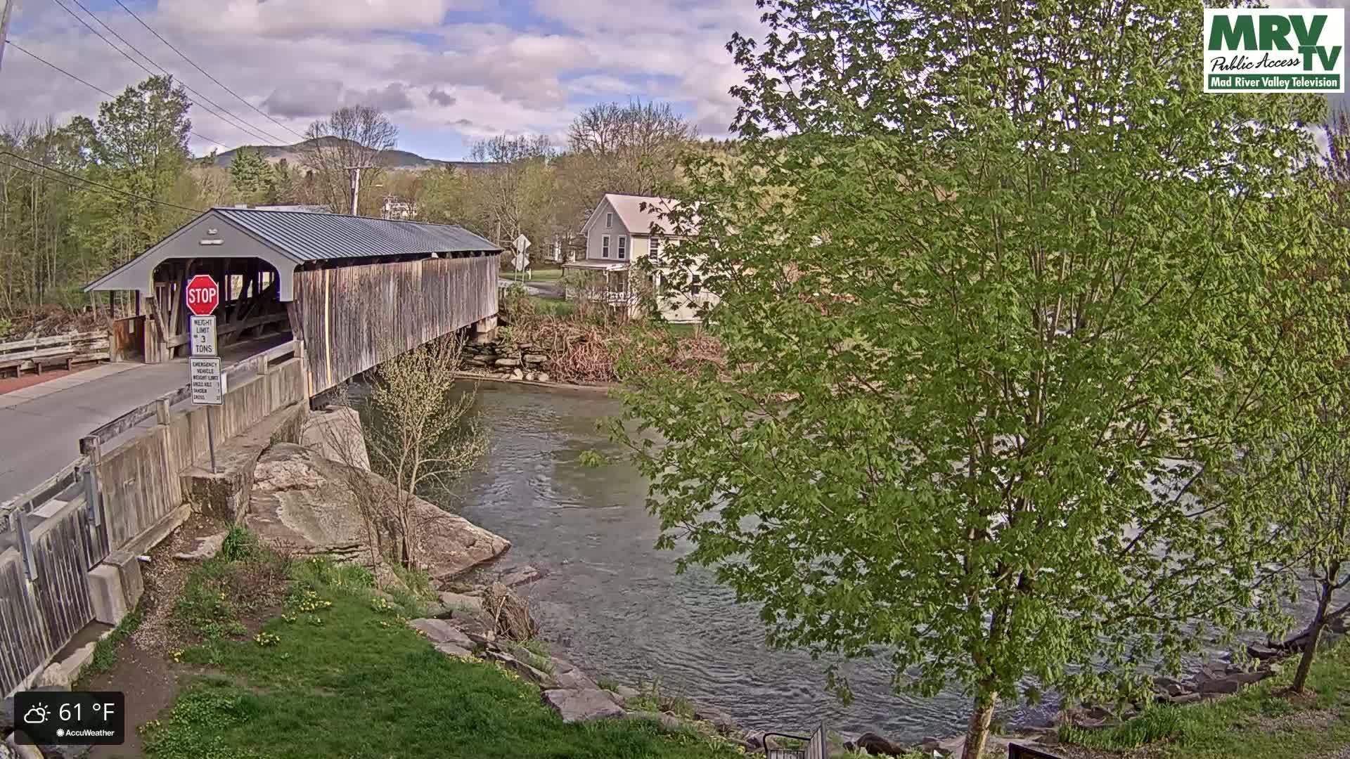 A covered bridge spans a river under a partly cloudy sky, with lush green trees and a house visible in the background.